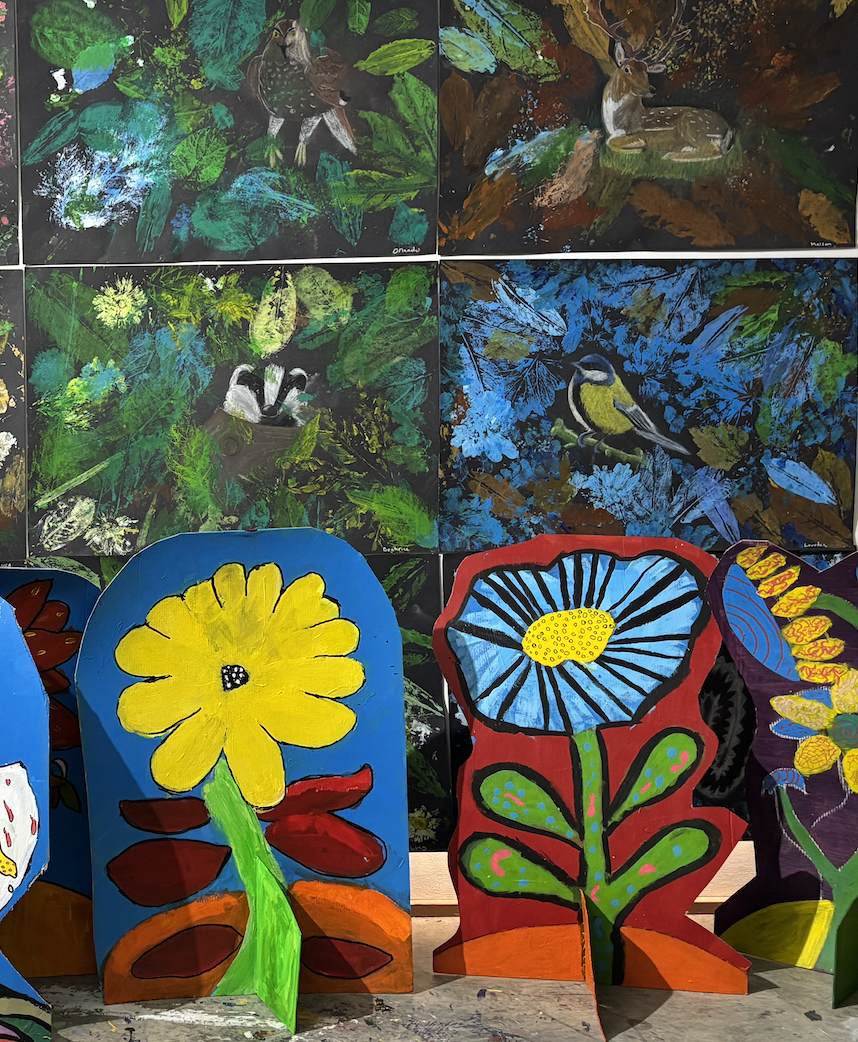 Colorful paintings and handcrafted art displays of birds, animals, and flowers on a wall and cut-out shapes on a table, with vibrant colors and detailed designs.