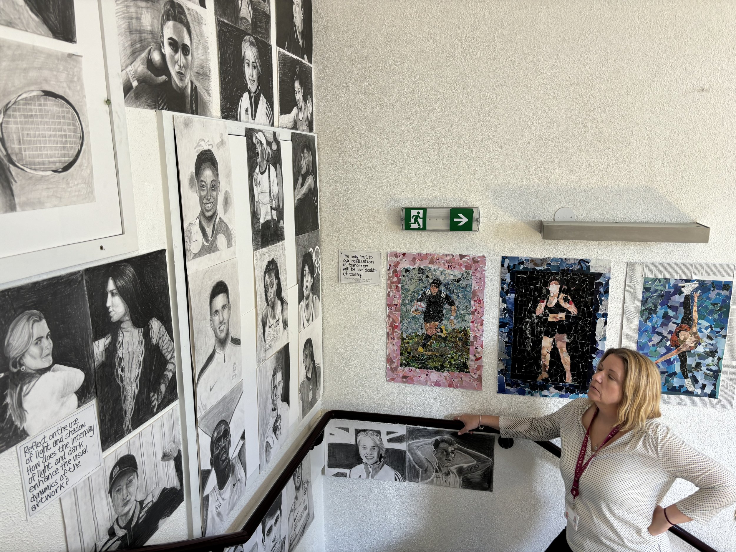 A woman leaning on a staircase railing in an art exhibit, with black and white portrait sketches on the left wall and colorful mosaic-style paintings on the right wall, including a soccer player, a boxer, and a performer.
