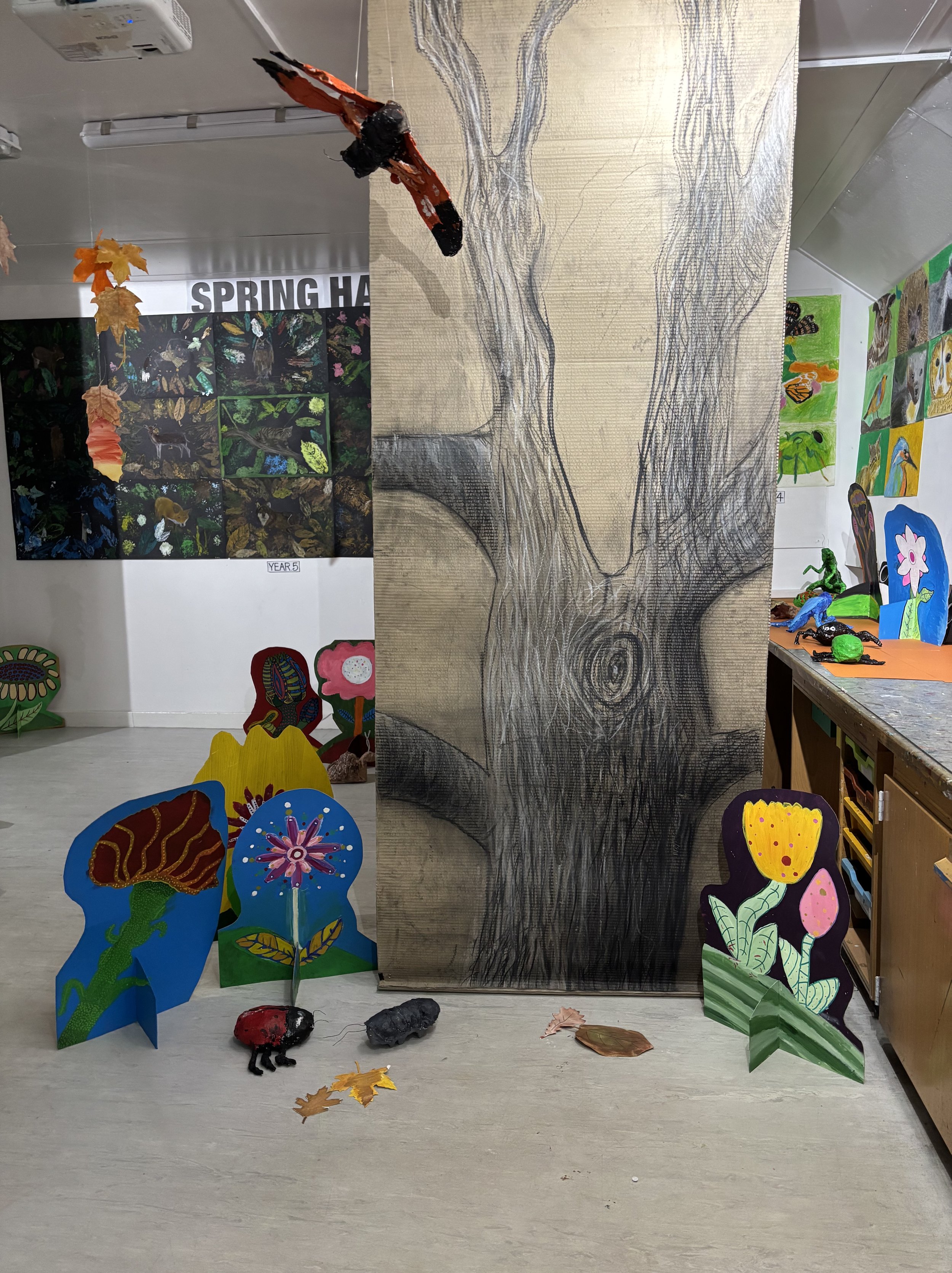 Indoor art display featuring painted and crafted nature-themed artworks, including a large sketch of a tree on a wooden panel, colorful cartoon flowers, insects, and animals, with some 3D butterfly and bug sculptures.