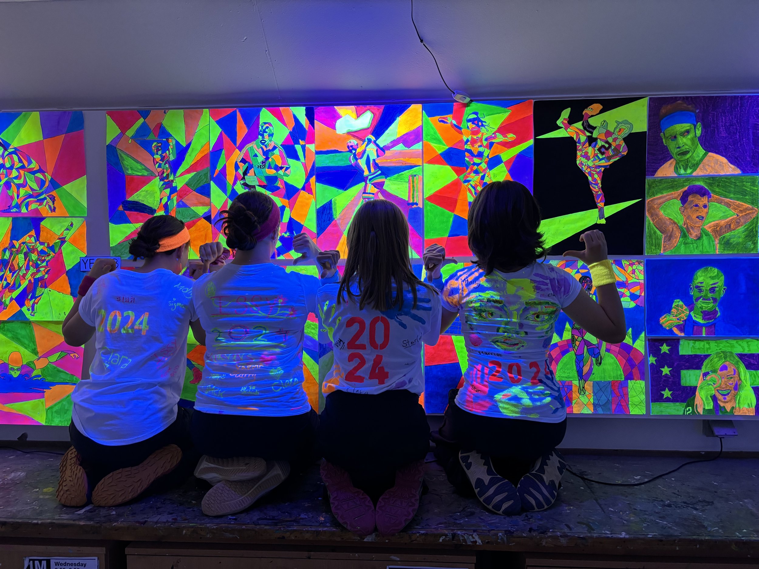 Four children kneeling and facing a colorful, glow-in-the-dark art display of athletes and portraits on a wall, with their backs to the camera.