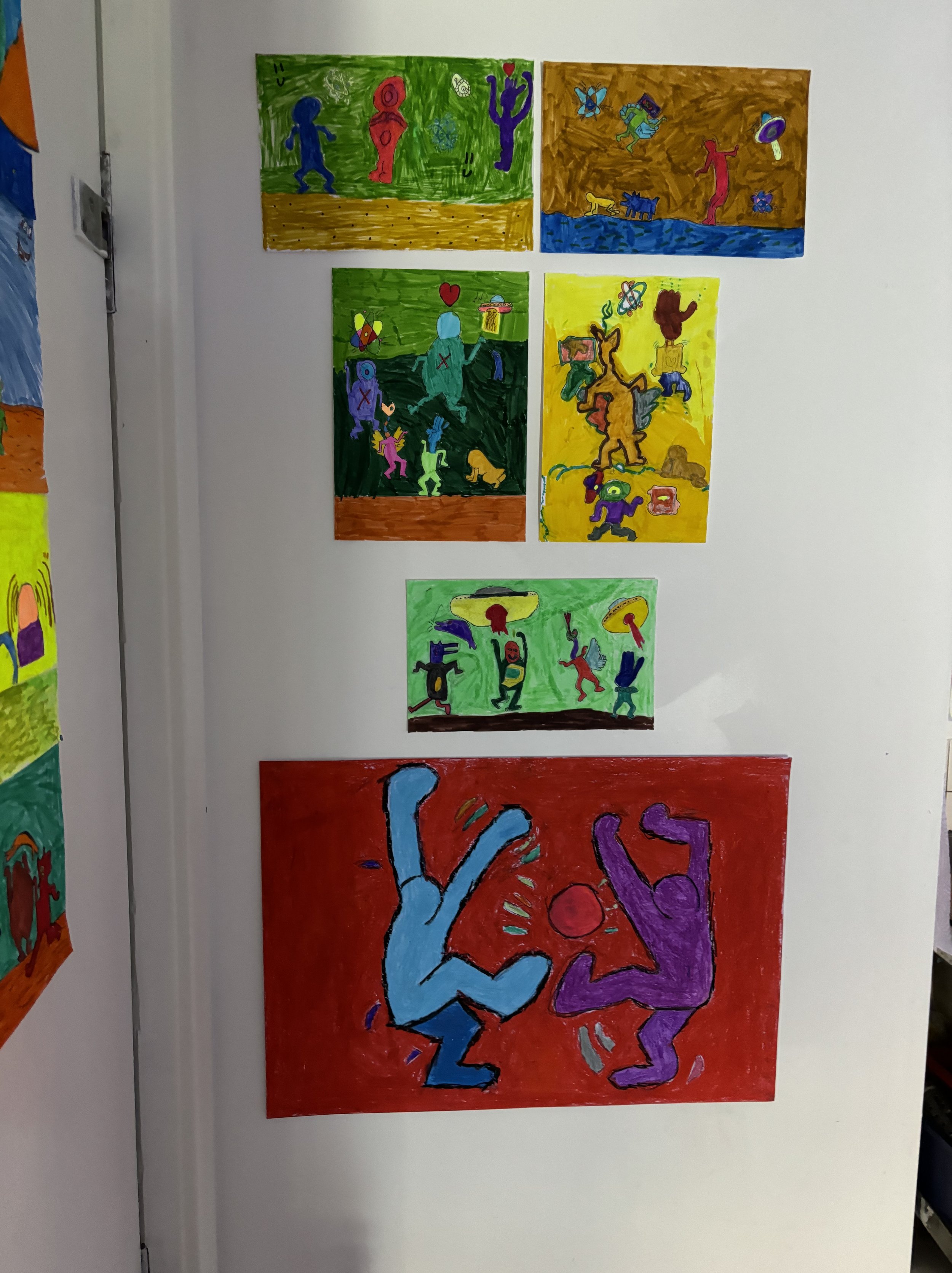 Colorful children's drawings of dancers and figures engaged in various activities, mounted on a white wall.