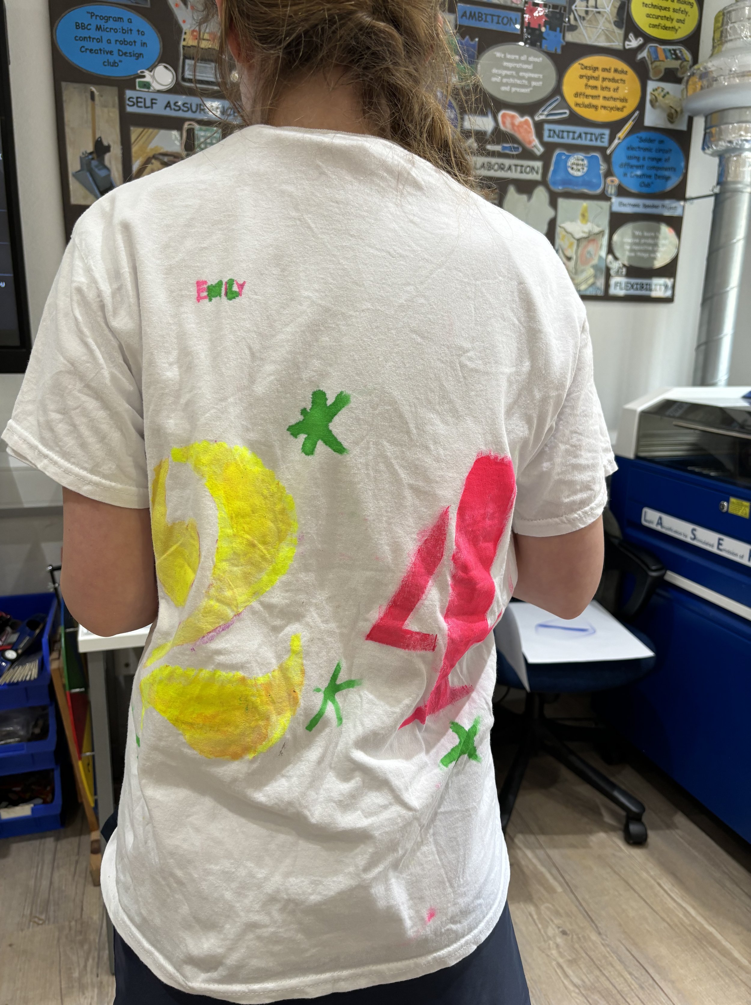 Back of a person wearing a white T-shirt with large painted numbers '24' in yellow and pink, with small green asterisks and 'EMILY' written in red and green near the top left.
