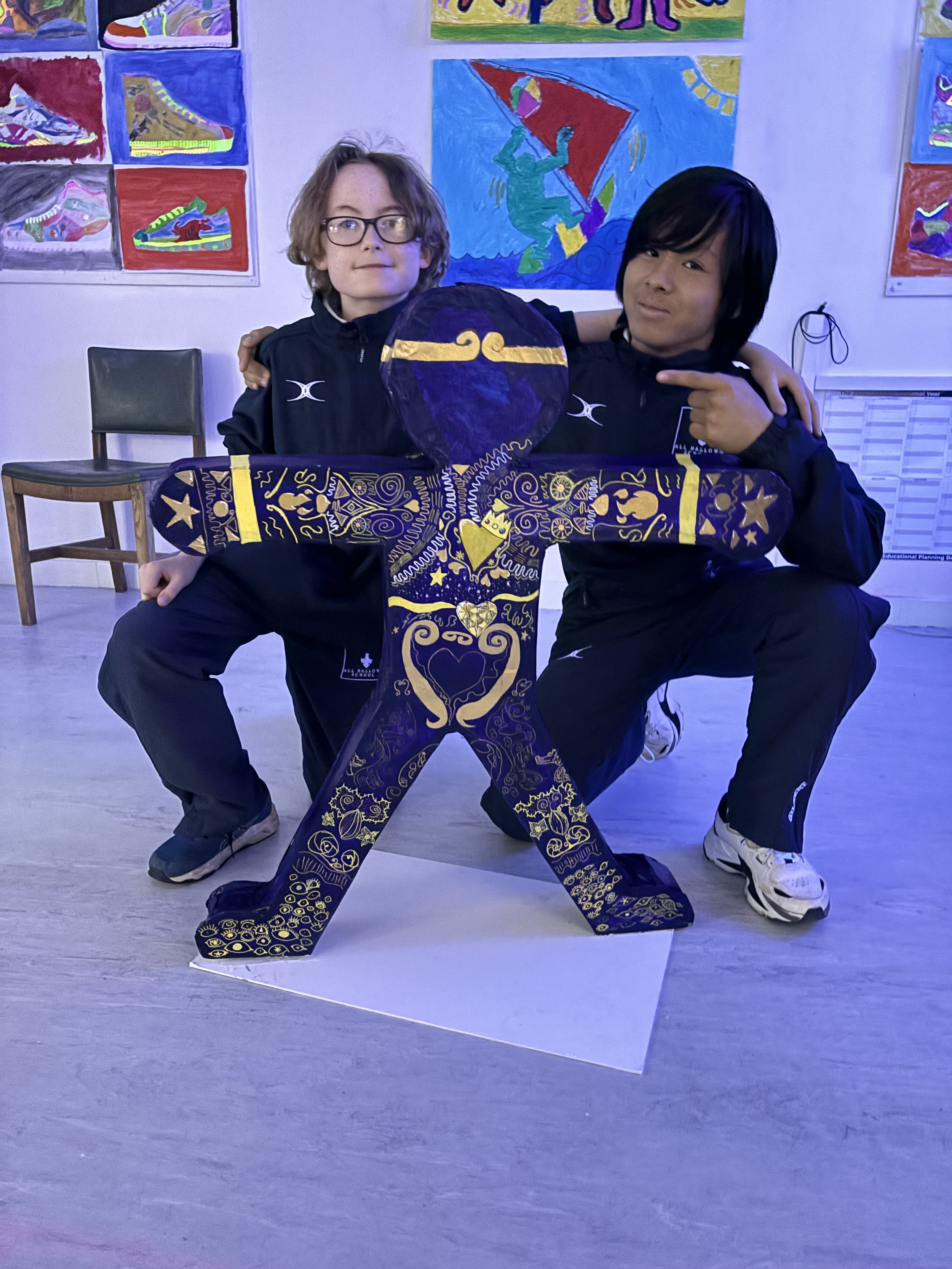 Two children posing with a decorated purple and gold cutout of a person holding a large medallion, in a room with colorful artwork on the walls.
