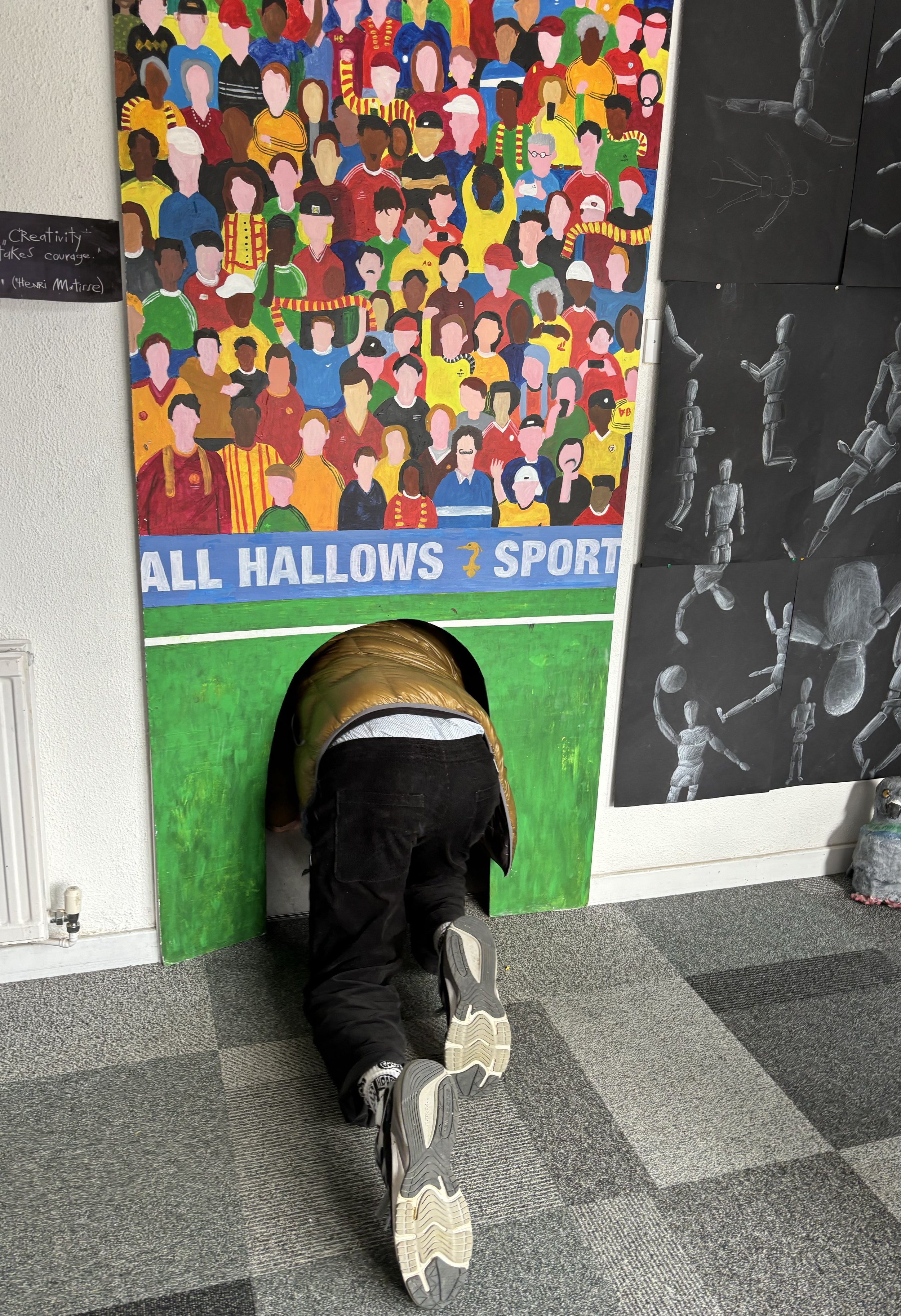 A person with a metallic gold jacket and black pants crawls through a small, circular opening in a colorful wall art piece depicting a crowd of diverse people. The wall art is segmented into a vibrant crowd on top and a green soccer field below with