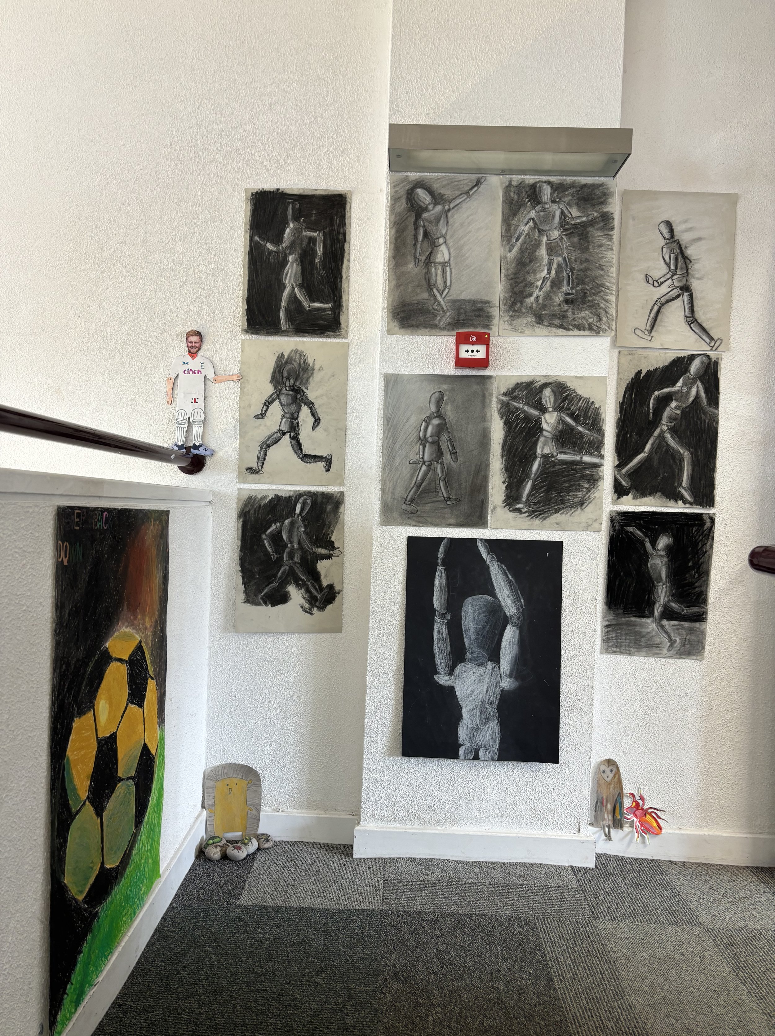 A white wall decorated with multiple black and white sketches of human figures in various poses. There is a small doll of a person in white with a beard on the upper left side, and a colorful painting of a soccer ball on the lower left. Two small fig