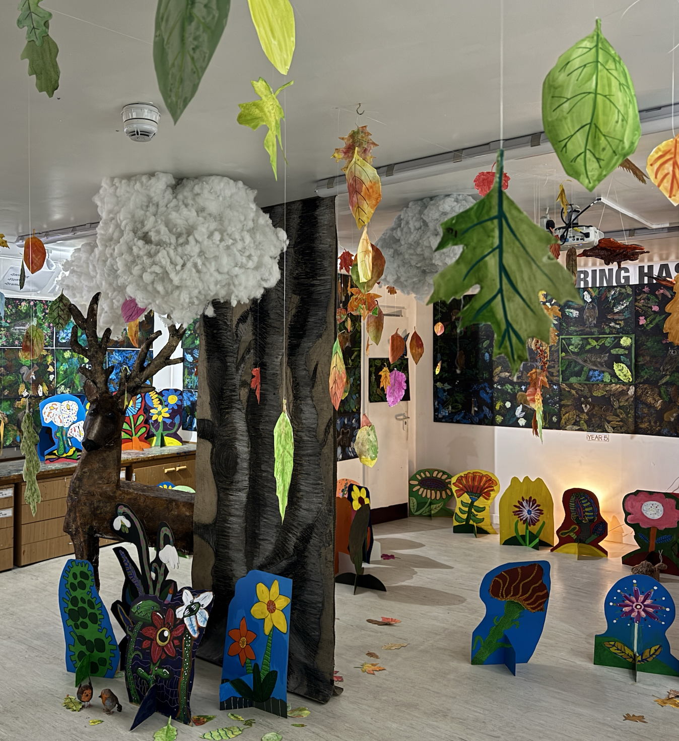 Indoor art display with a painted cardboard tree, paper clouds, colorful cutouts of flowers and insects, hanging leaves, and a wood sculpture of a deer. Decor includes painted panels of nature scenes and autumn leaves scattered on the floor.