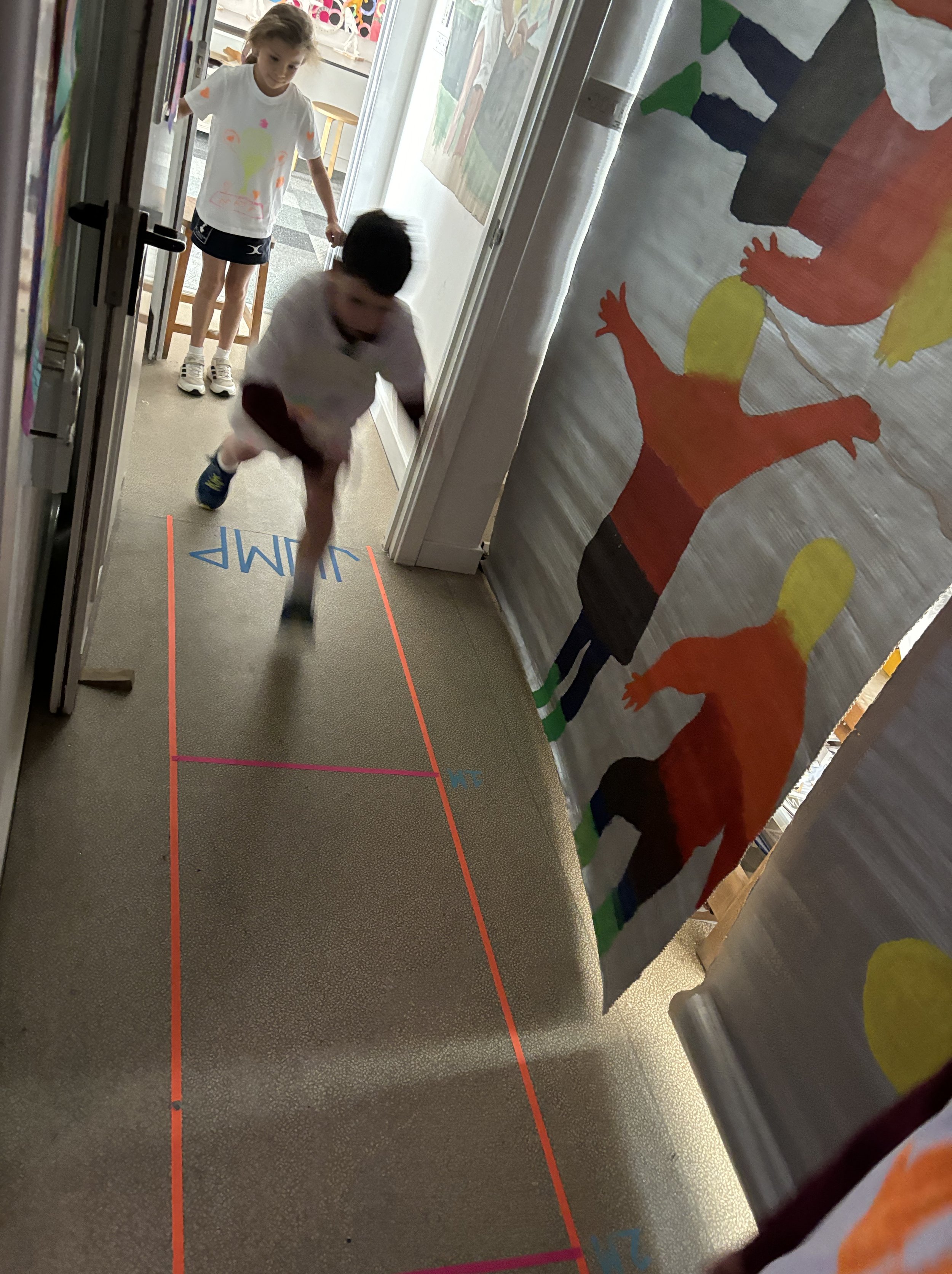 Two children playing a game of hopscotch on the hallway floor, marked with colored lines and numbers, next to colorful animal artwork on the wall.