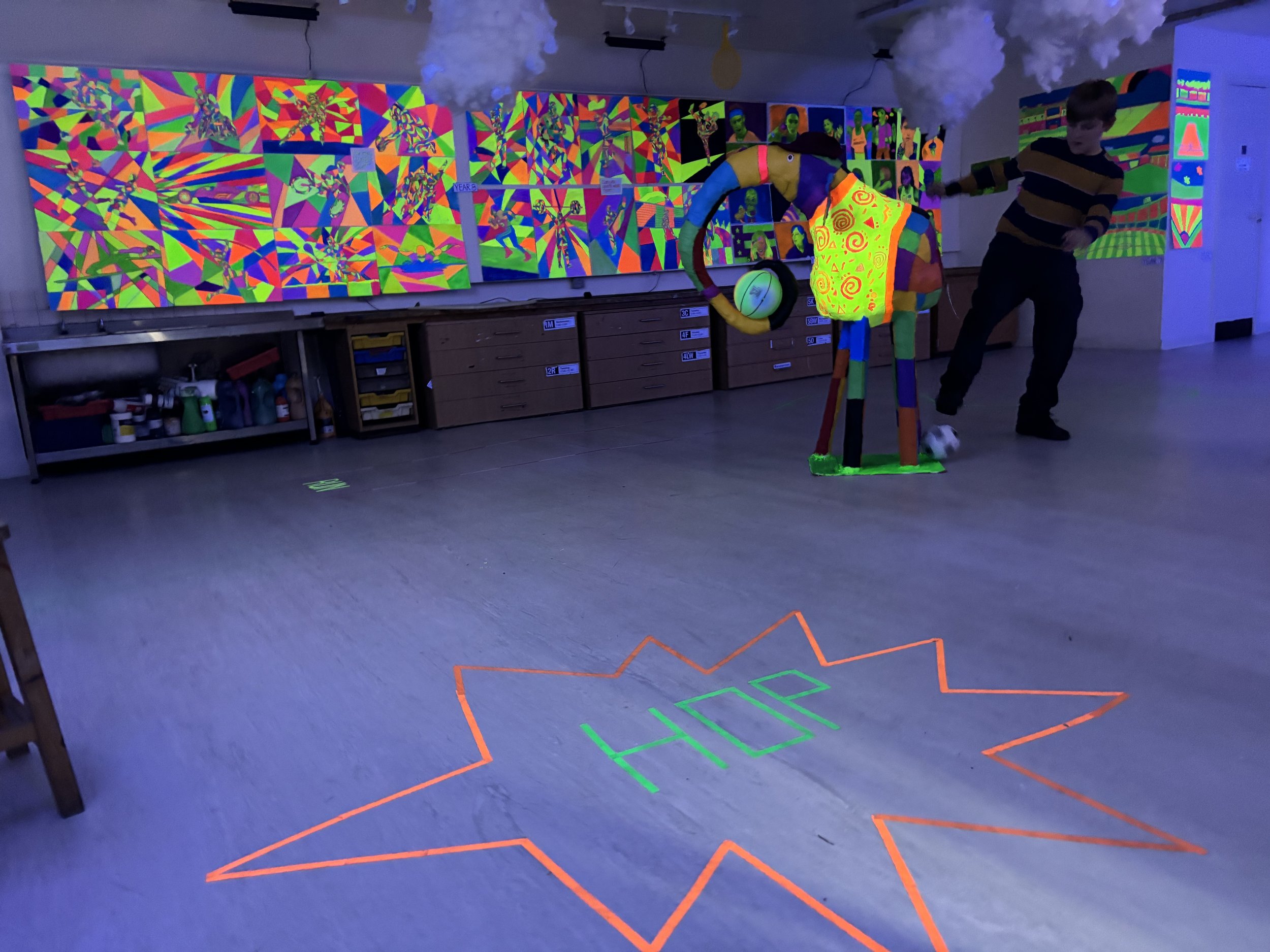 Indoor room decorated with neon and glow-in-the-dark art, featuring a large painted mural on the back wall, a neon star with the word 'HOP' on the floor, a colorful foam sculpture of headphones and a figure, and a person playing soccer in the glow-in