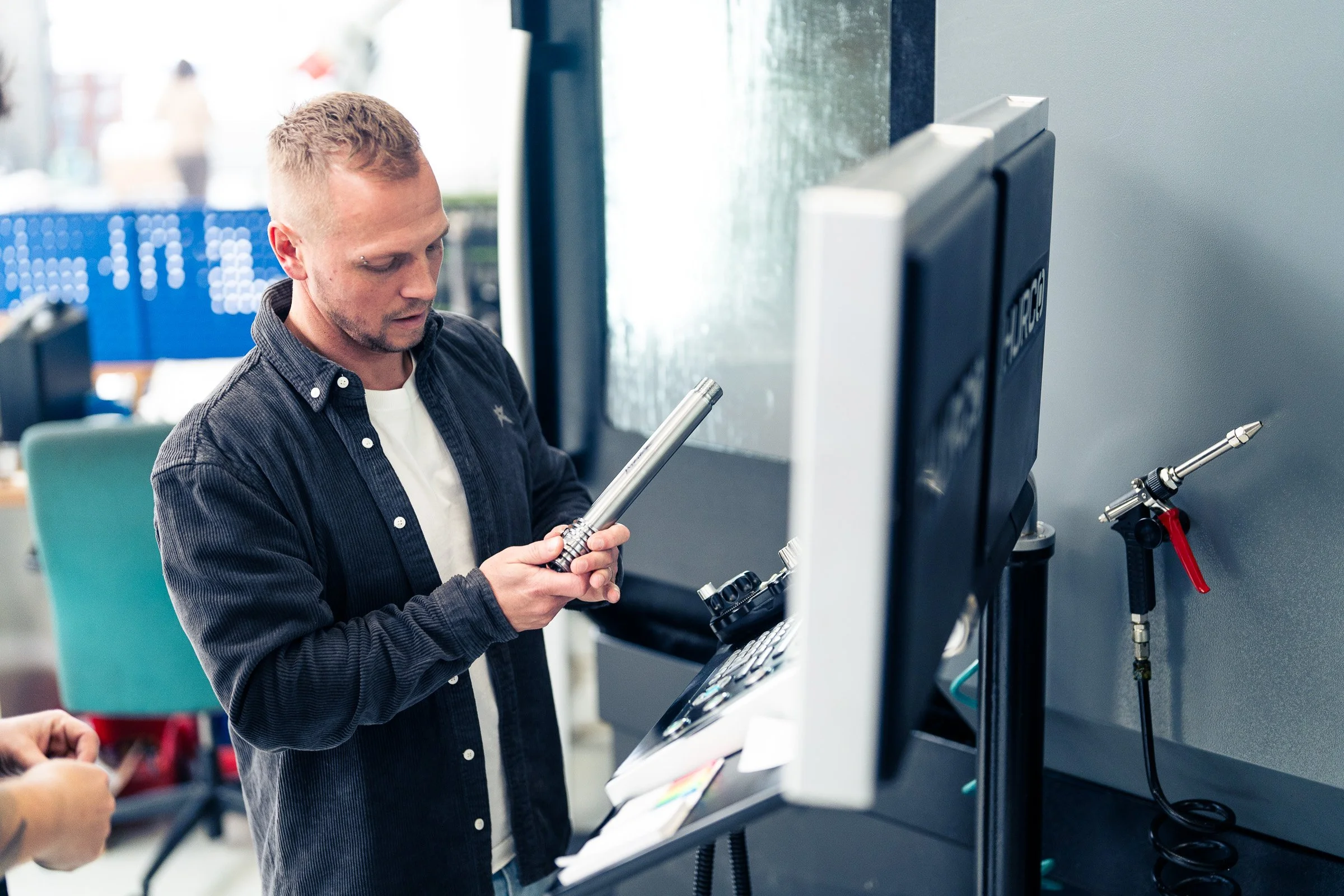 Ein Mann mit Kurzhaar und Bart, der in einem technischen oder wissenschaftlichen Umfeld an einem Gerät arbeitet, hält ein metallisches Werkzeug in der Hand.