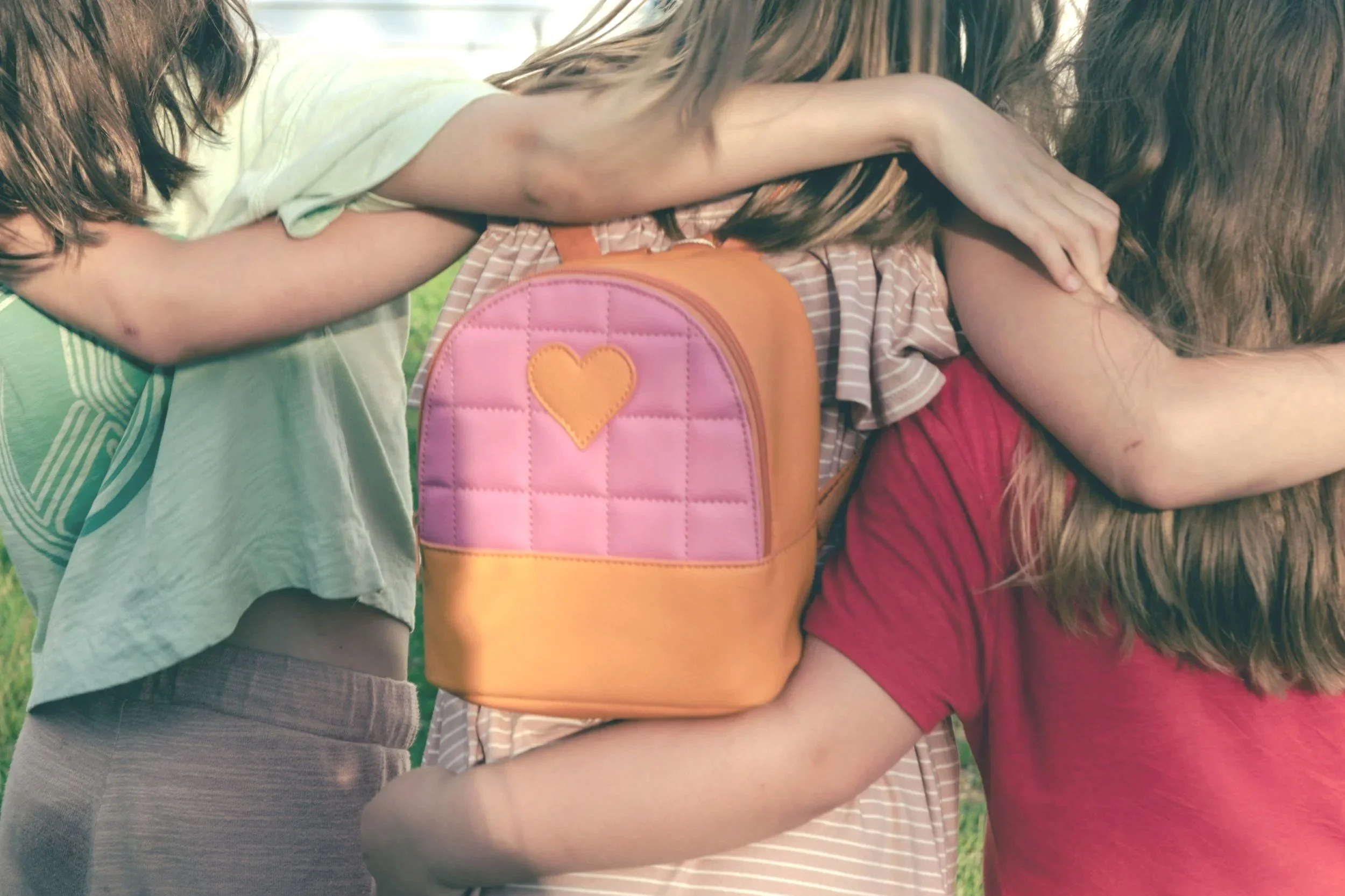 Three children hugging outdoors, one girl wearing a pink backpack with a yellow heart, two girls in striped and red shirts.