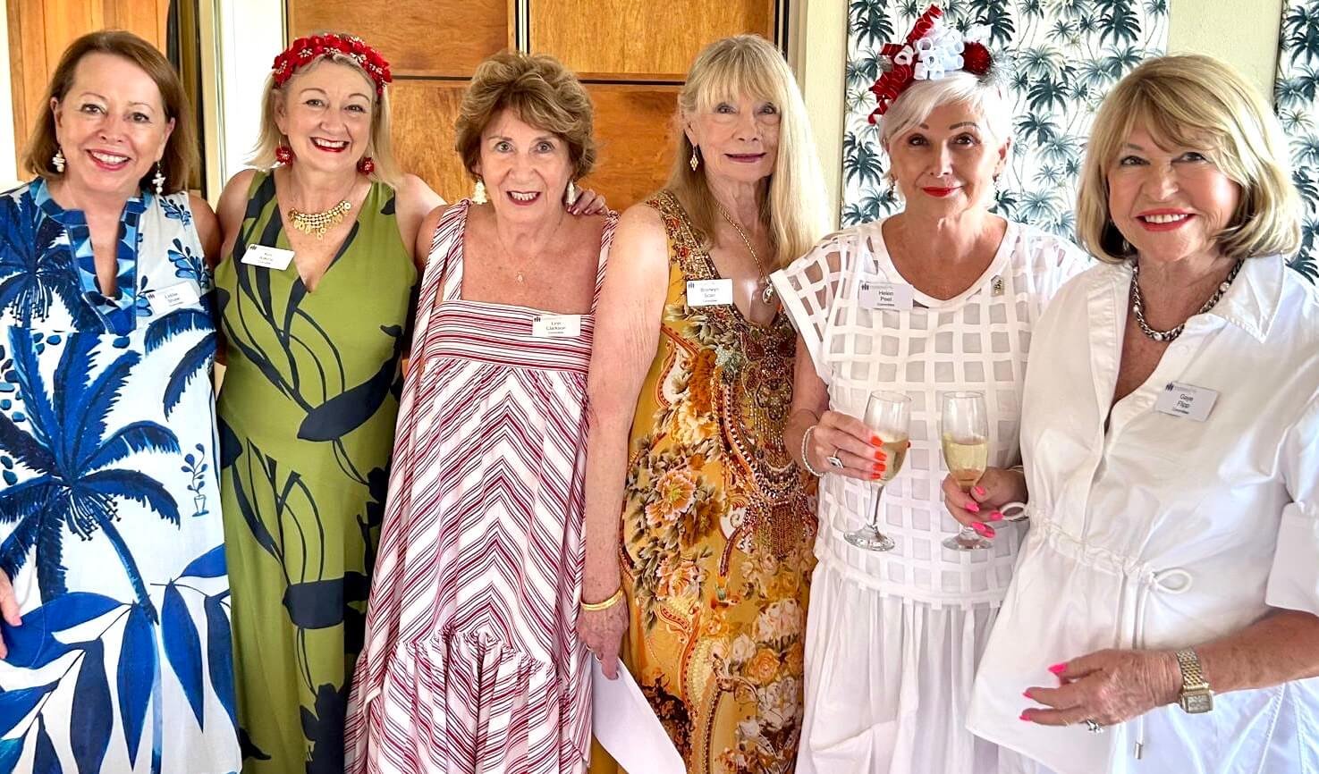 A group of seven women standing together at a social gathering, some holding glasses of white wine, inside a room with tropical-themed wallpaper.