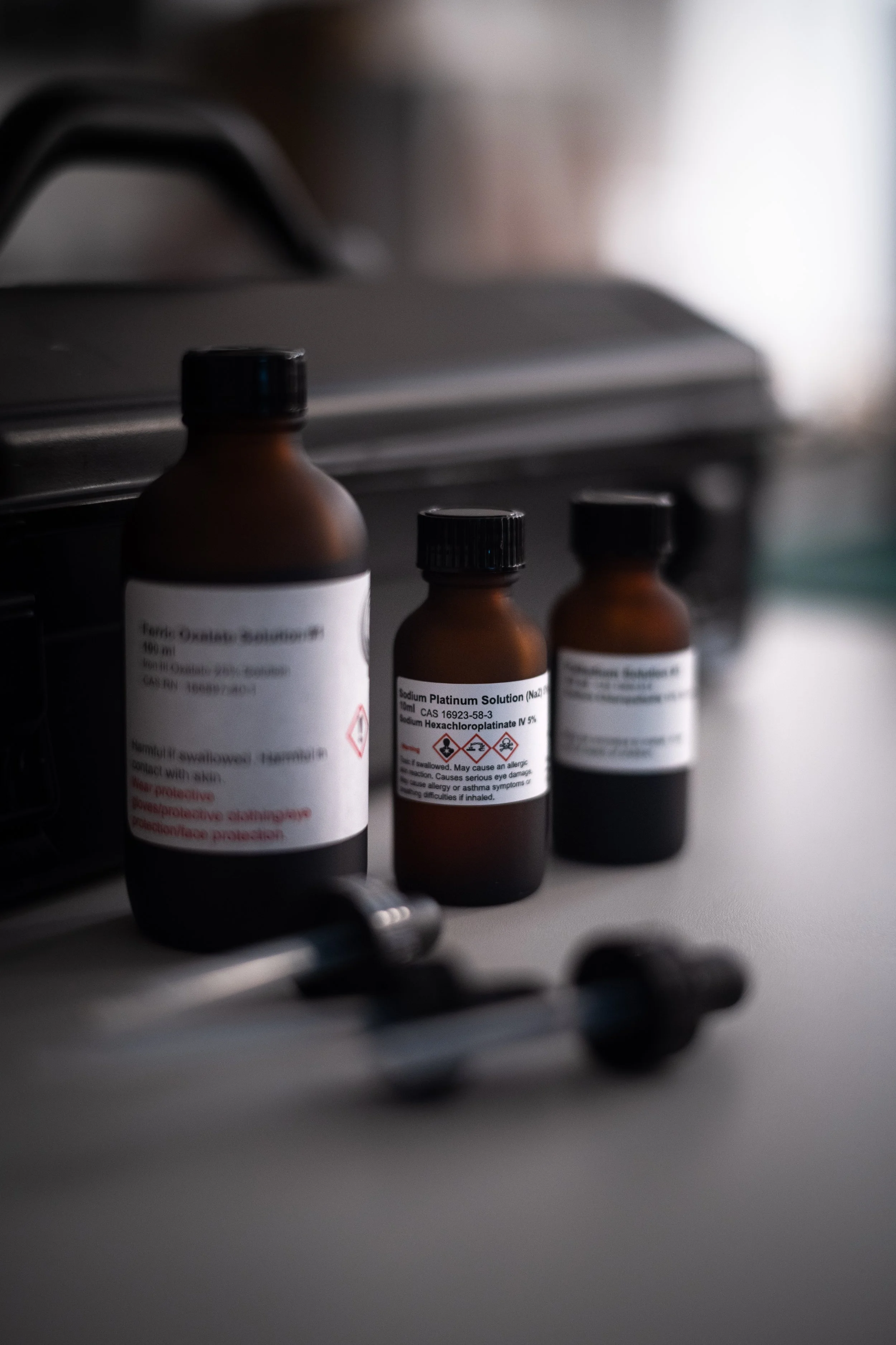 Three brown bottles with medical labels, possibly for medications or chemicals, placed on a white surface with a pair of metal forceps nearby.