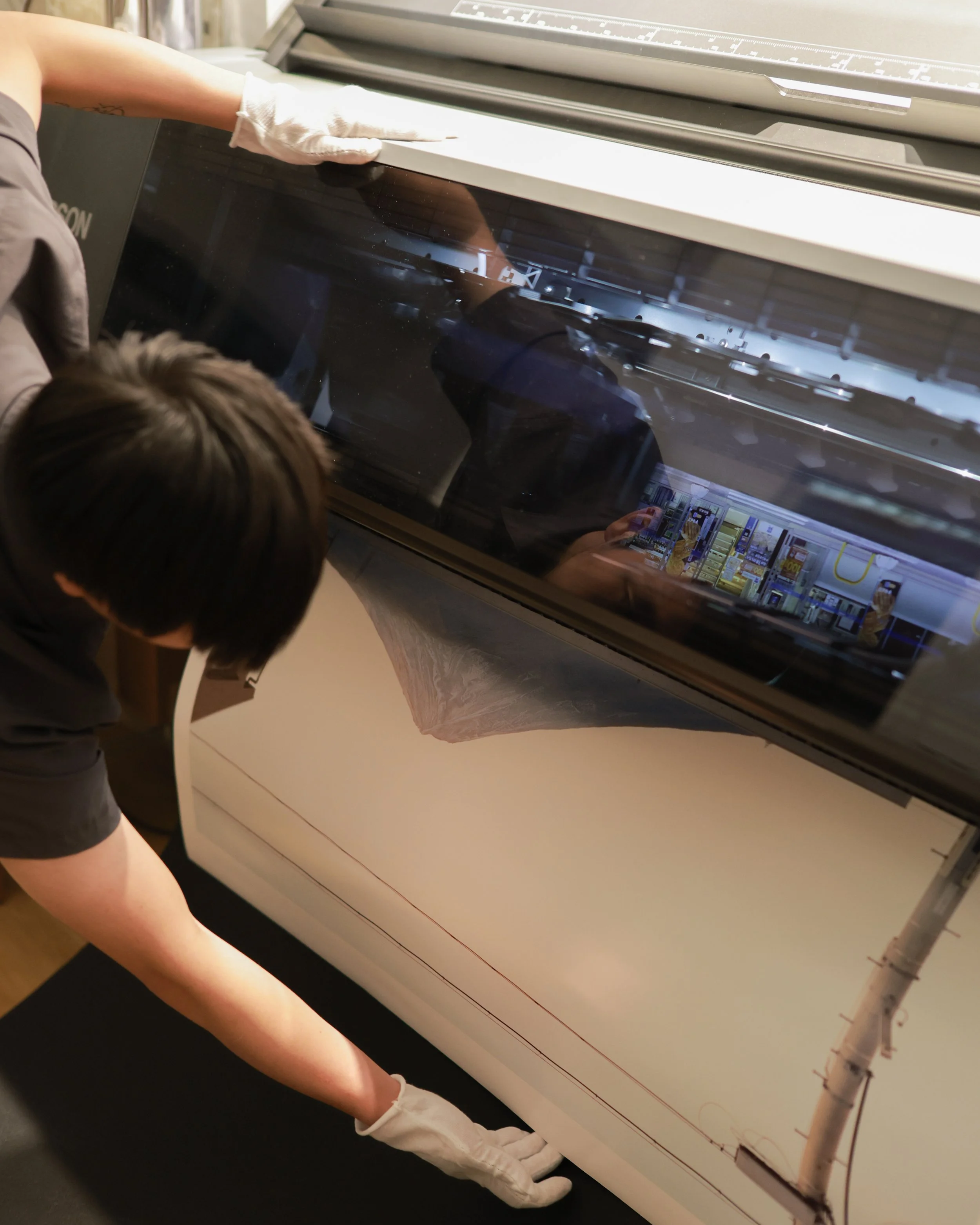 A person using a large machine, possibly a flatbed printer or scanner, wearing white gloves and leaning over the machine, with a reflection of their hands and part of their face visible on the glass.