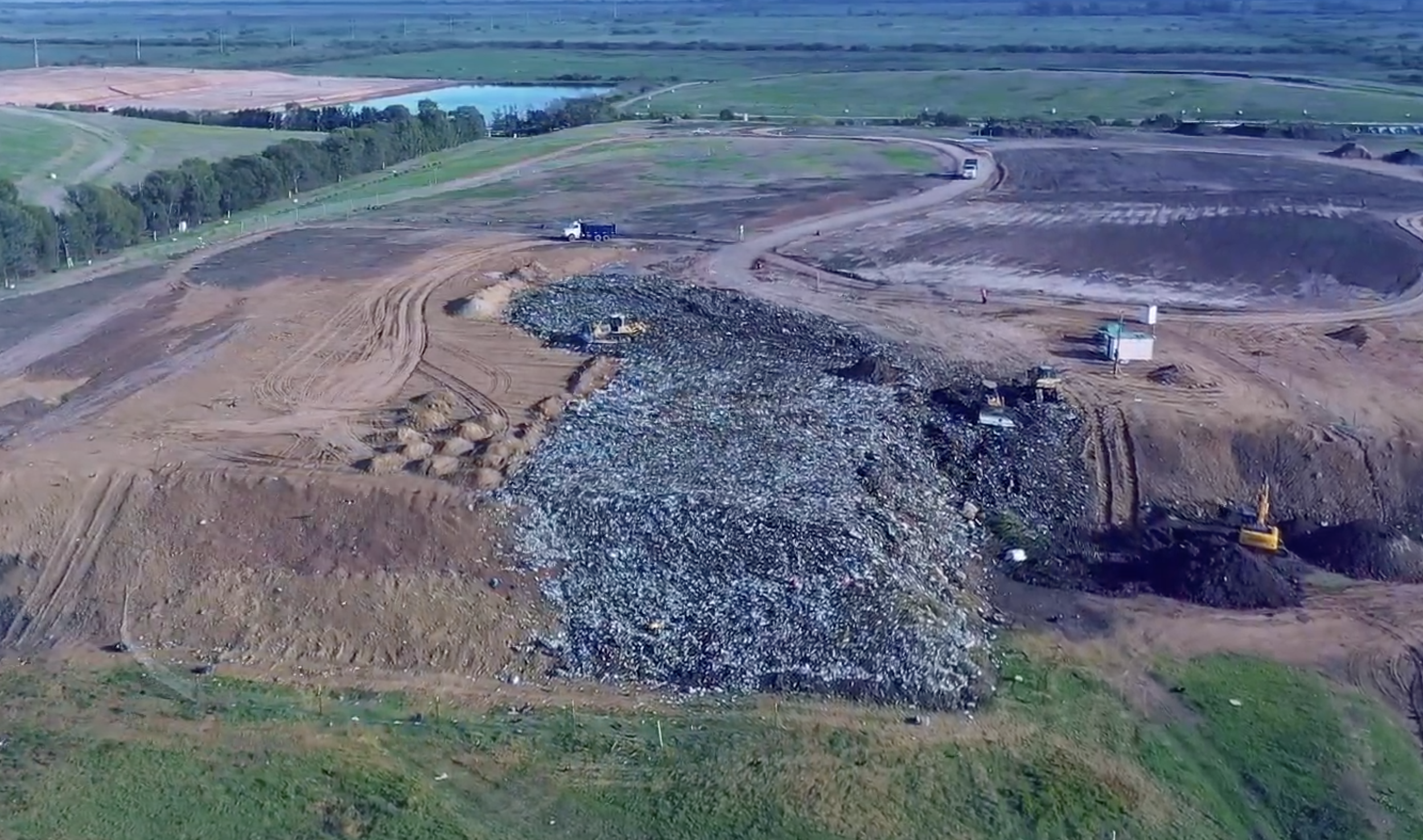 Zona de construcción con maquinaria y tierra removida, cerca de un cuerpo de agua y áreas verdes.