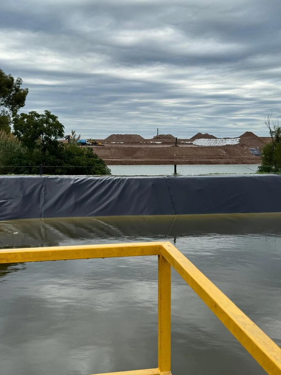 Imagen de un área de construcción junto a un cuerpo de agua, con montículos de tierra, maquinaria y una estructura de bloques de concreto, debajo de un cielo nublado.