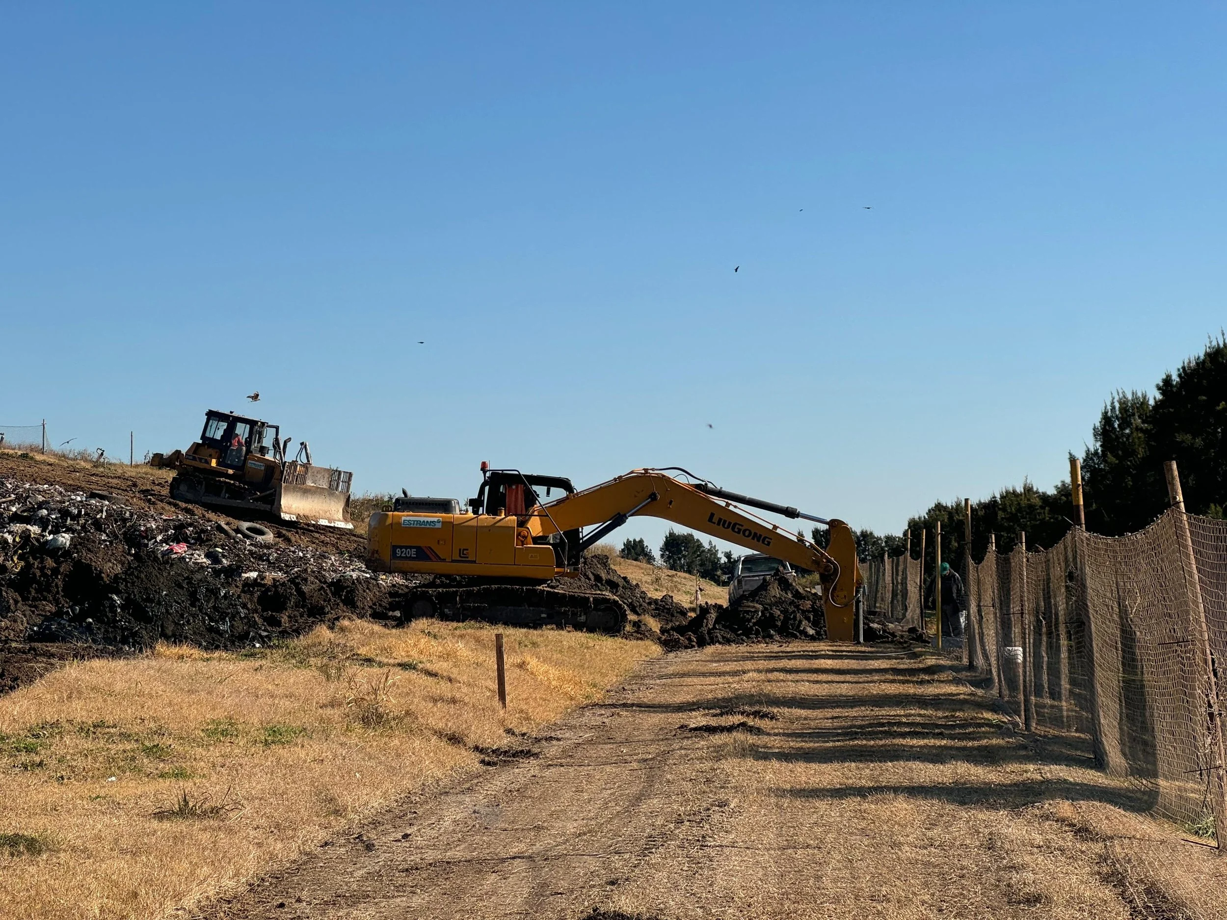 Máquinas excavadoras trabajando en un sitio de construcción de tierra y rocas, con un cerco de fibra de vidrio en un lado y césped seco en primer plano, bajo un cielo despejado.