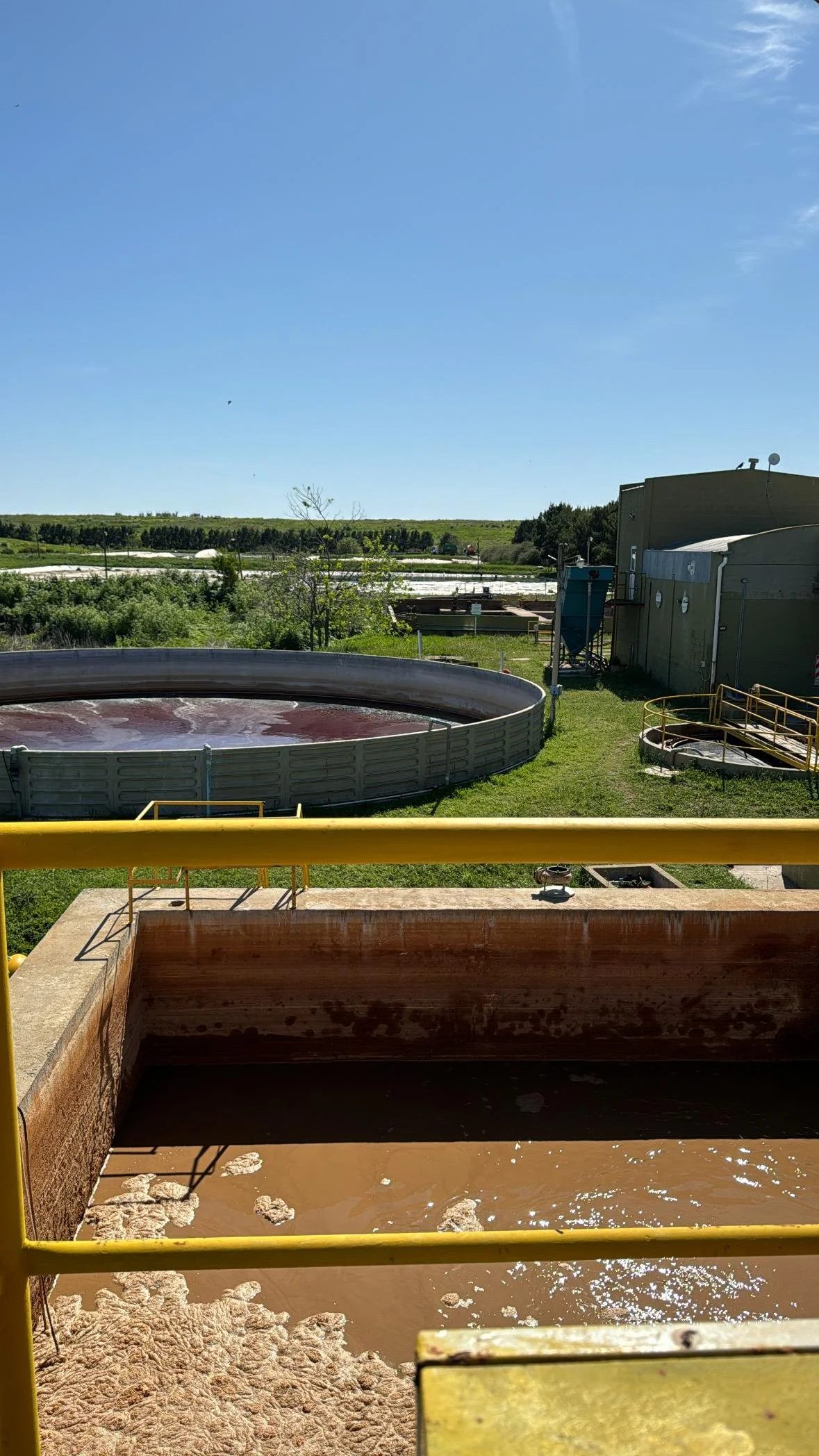 Tanque de agua marrón en primer plano con rampa amarilla, más atrás un estanque circular con agua de color rojizo, y un paisaje de campo con vegetación y un cielo despejado.