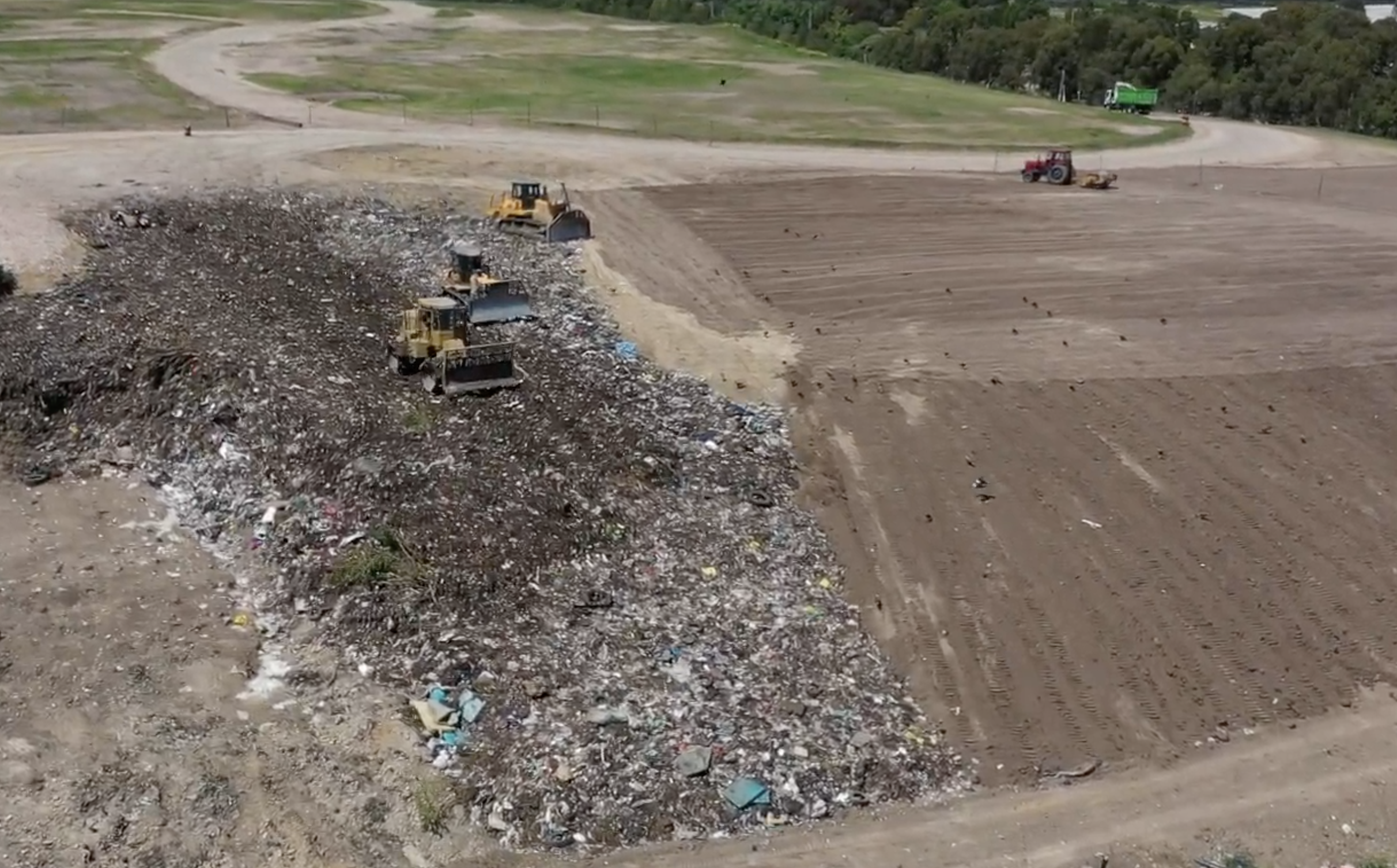 Vista aérea de un vertedero con maquinaria pesada que recoge basura, en un terreno con partes de suelo despejado y áreas de tierra arada, rodeado de vegetación y árboles al fondo.