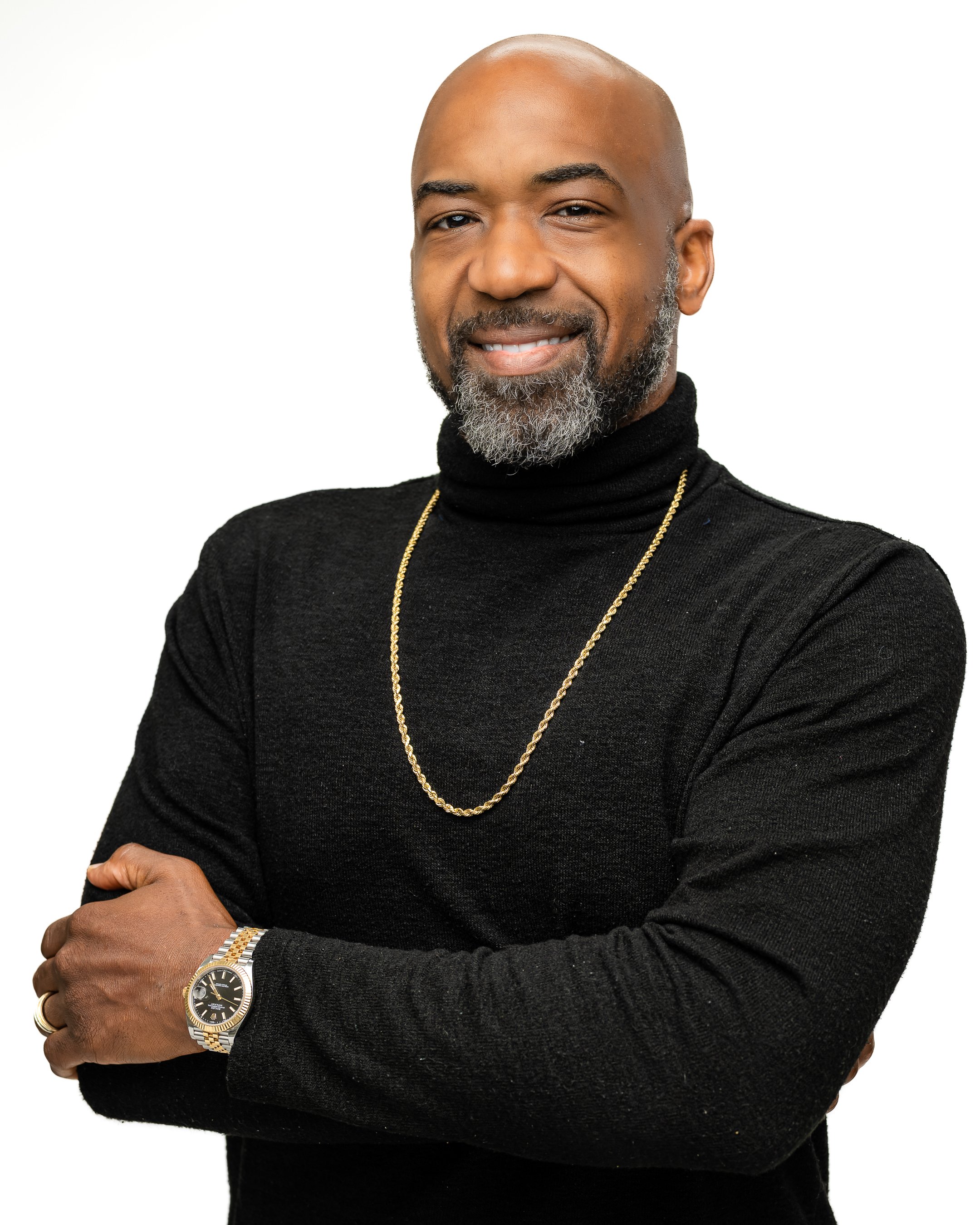 A confident man with a beard and bald head, smiling, wearing a black turtleneck sweater, gold chain, and a silver and gold watch, posing with arms crossed against a white background.