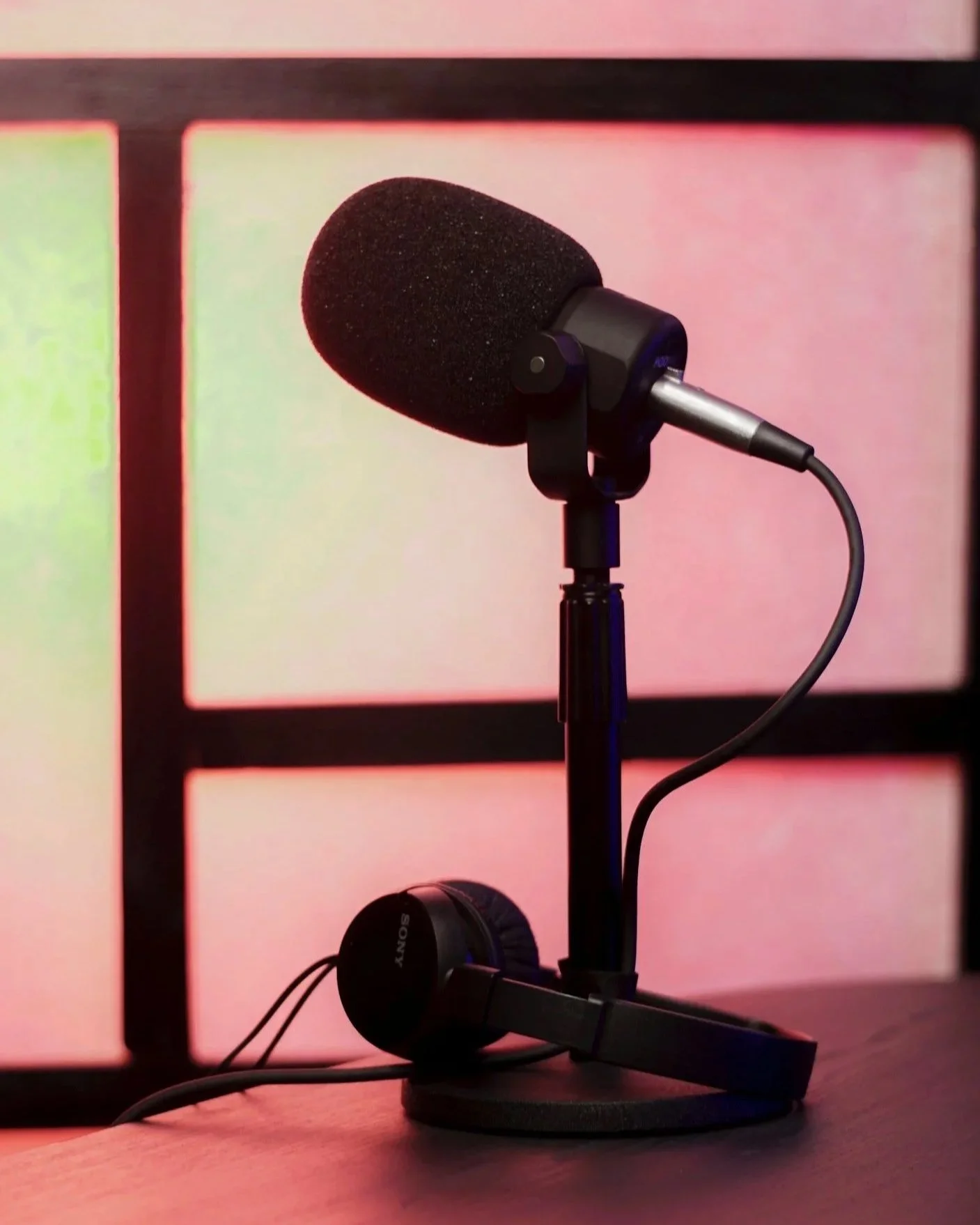 A microphone mounted on a stand with headphones resting on the desk in front of it, in front of a colorful blurred background.