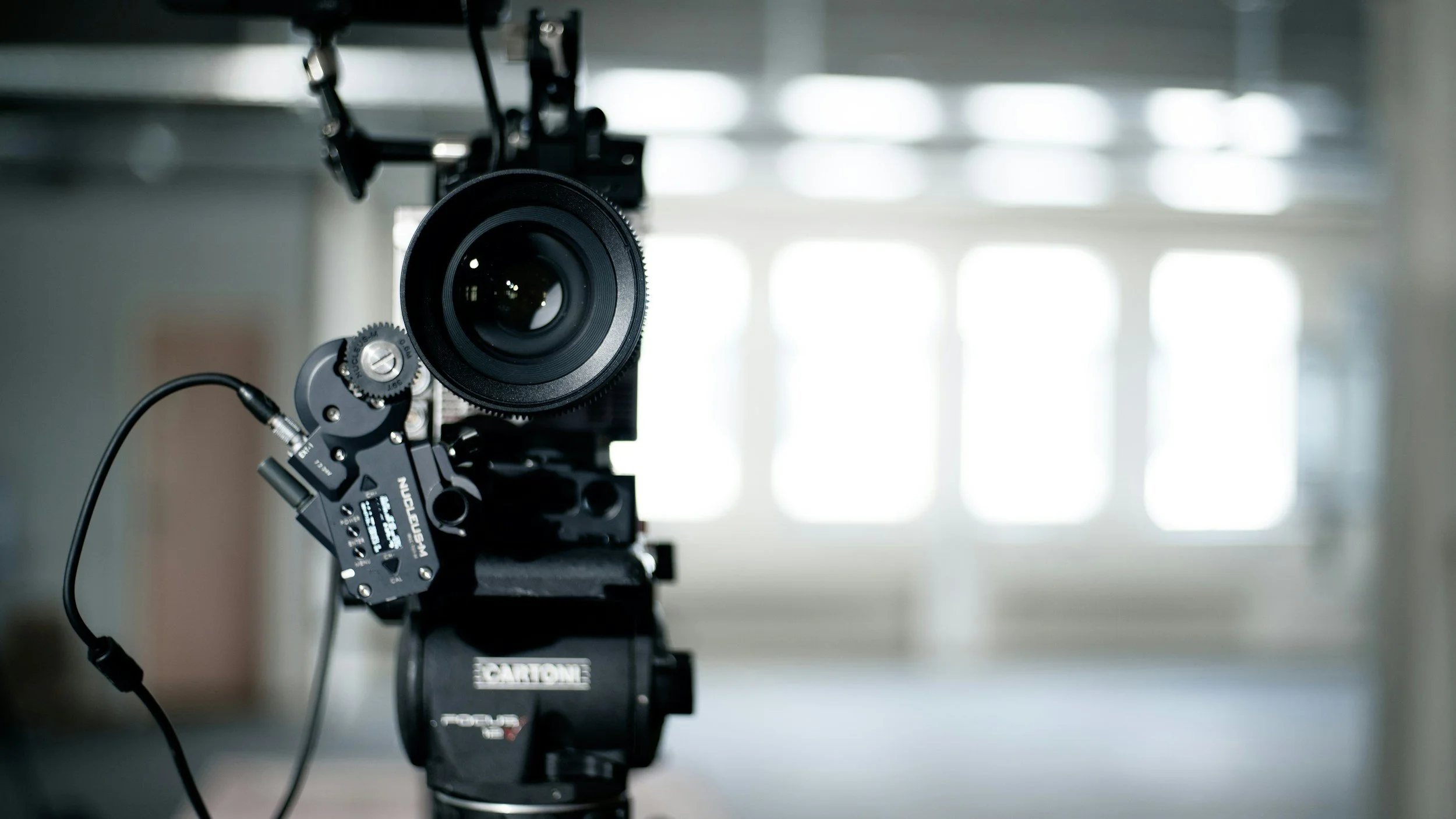 Close-up of a professional camera mounted on a stabilization rig, with a large lens and various adjustment components, in an indoor studio setting with bright natural light.