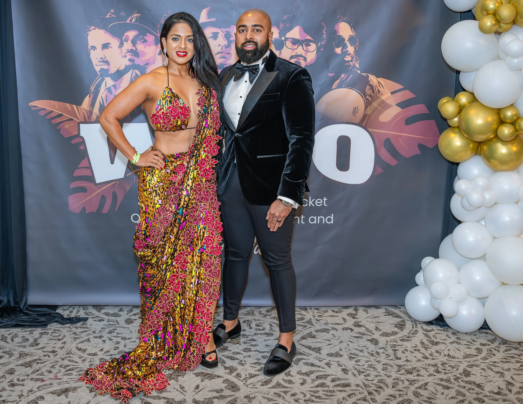 A man and woman dressed in formal attire posing together at an event with a backdrop and balloon decorations.