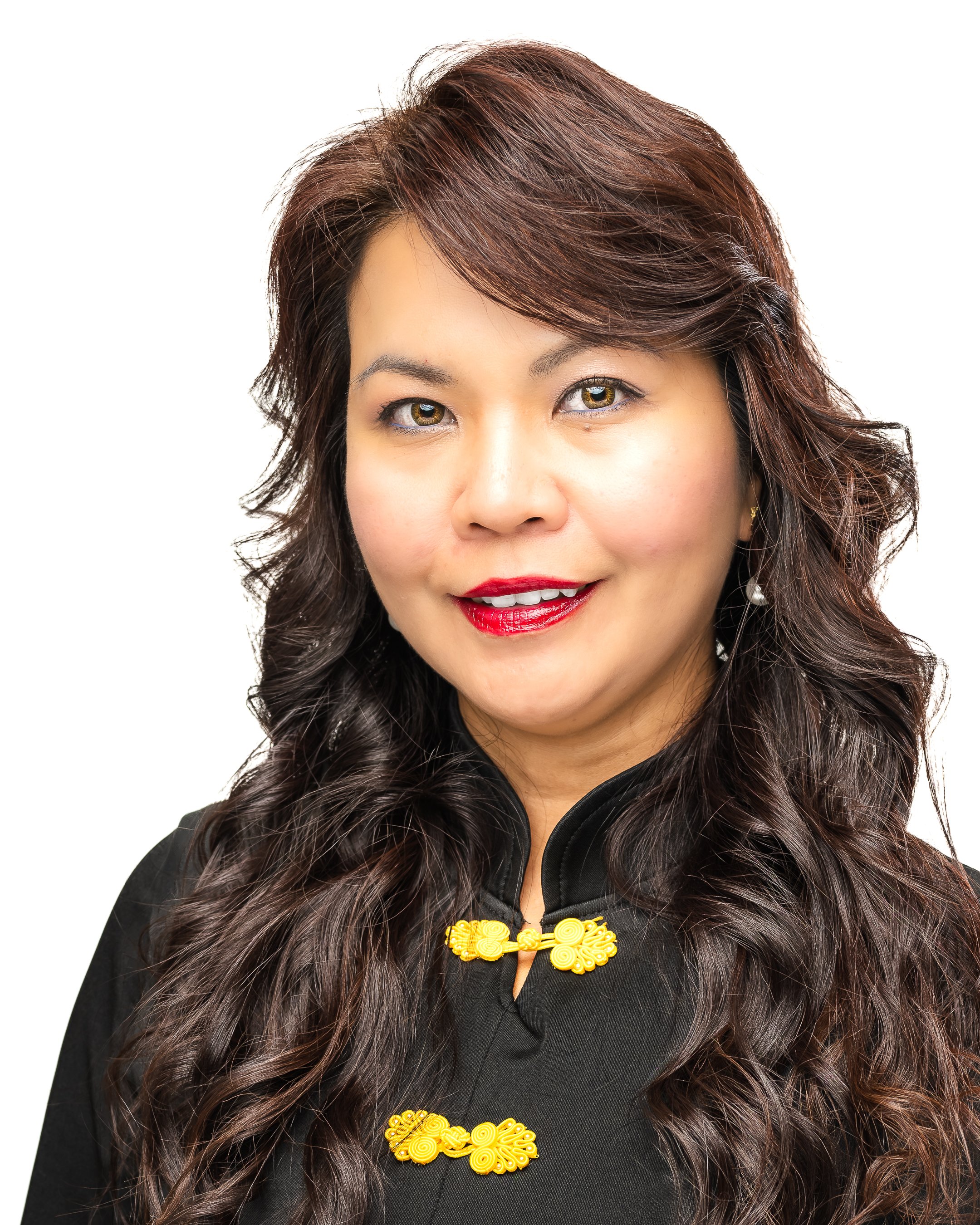 Portrait of a woman with long, dark, wavy hair, wearing a black jacket with yellow decorative buttons, red lipstick, and earrings, smiling against a white background.