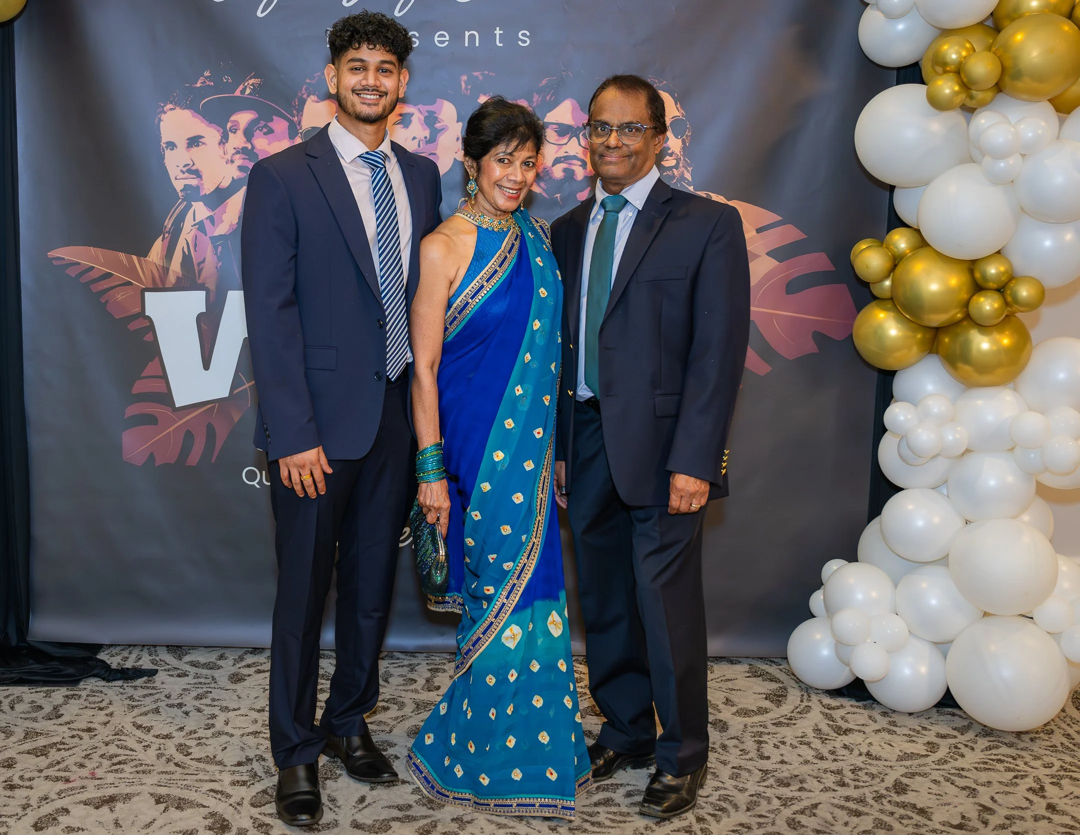 Three men and one woman dressed in formal attire standing together at an event with a backdrop and a balloon decoration to the right.