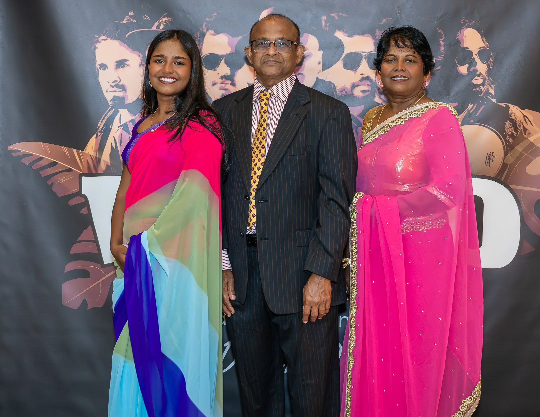 A family of three standing together in front of a dark backdrop with colorful graphics. The woman on the left is wearing a multicolored saree with shades of pink, green, blue, and purple. The man in the middle is dressed in a dark pinstripe suit with