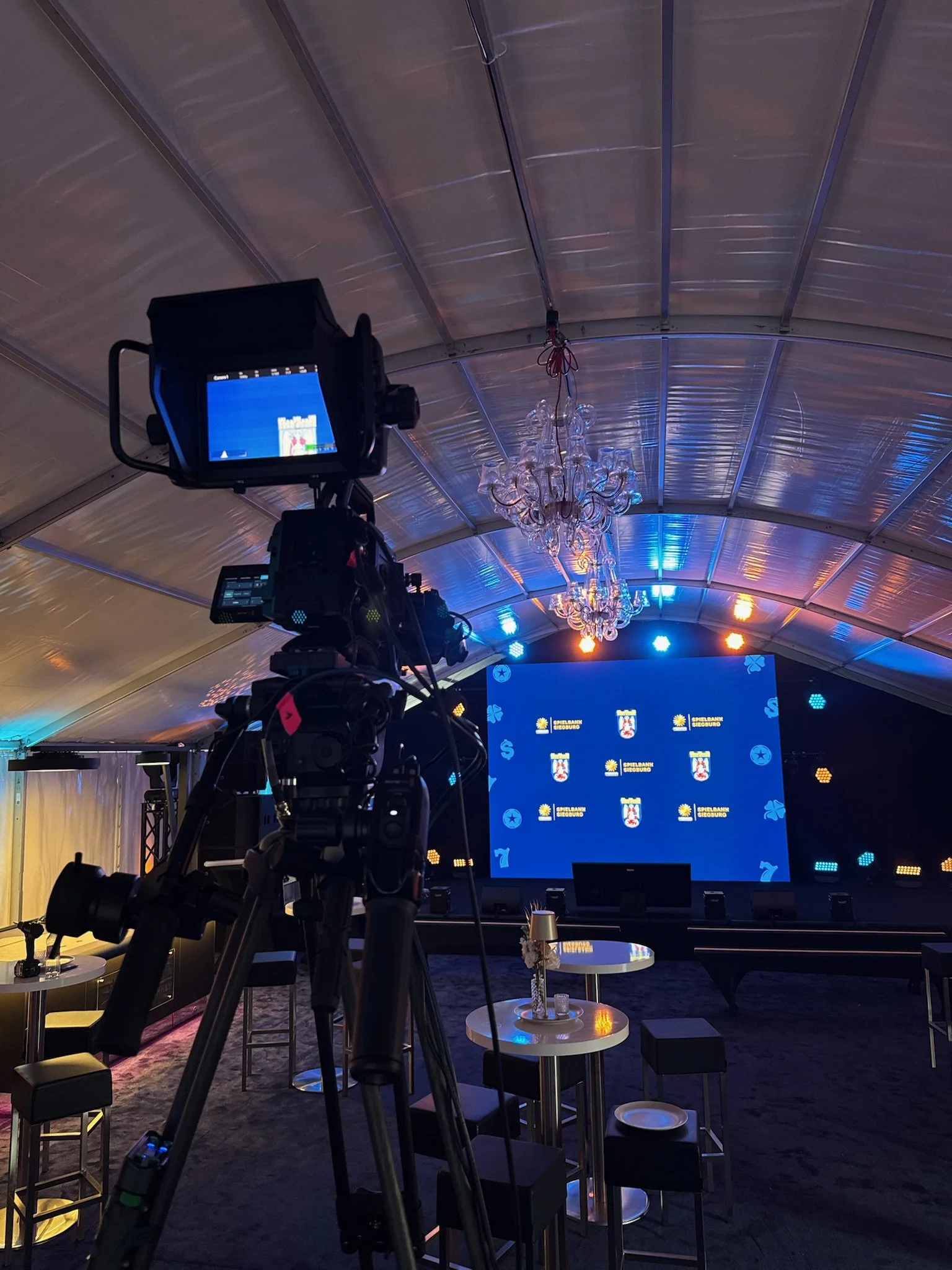 Ein Kamerastativ vor einer großen Leinwand in einem festlich dekorierten Zelt, mit Tischen, Stühlen und einer Kronleuchterdecke.
