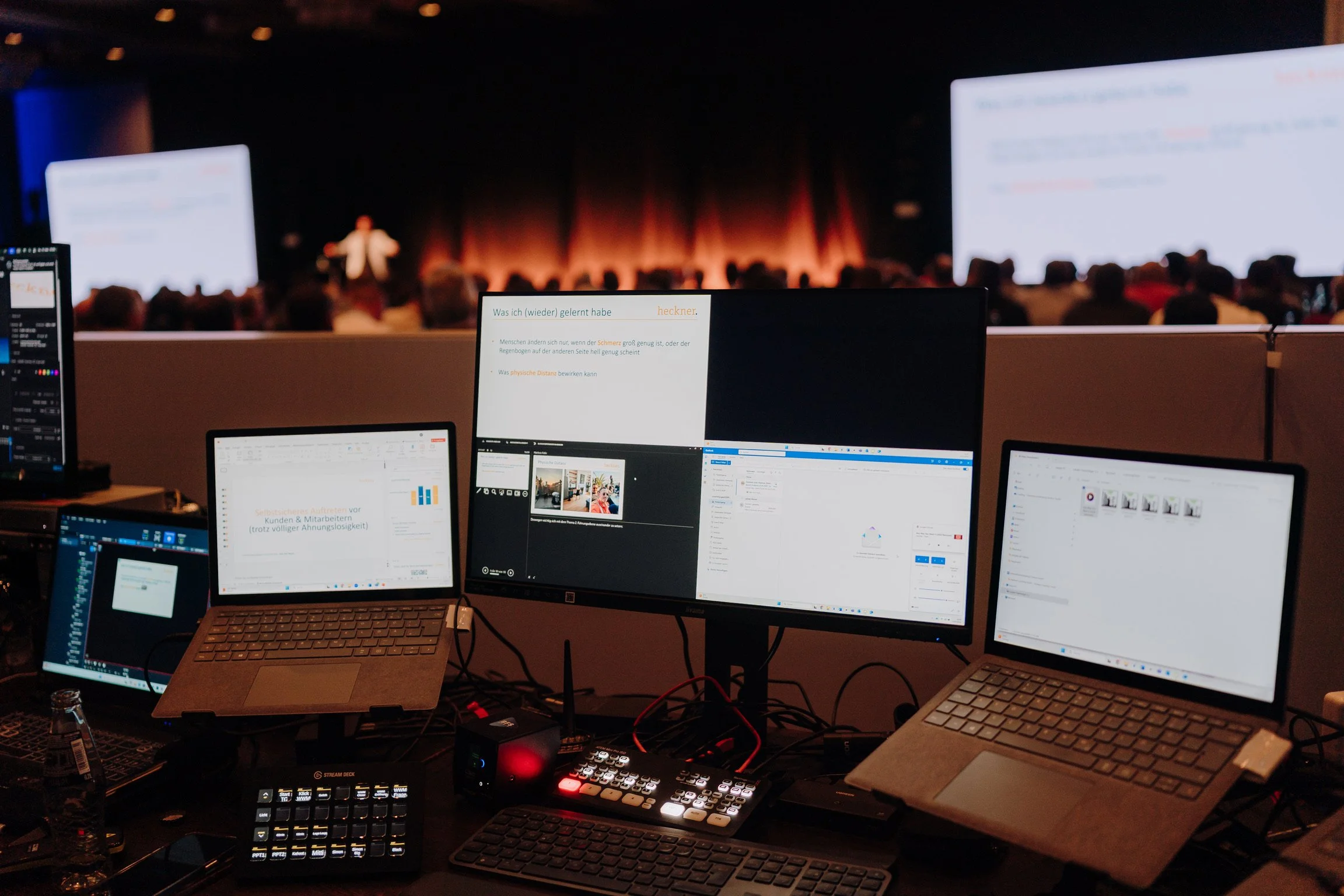 Mehrere Computerbildschirme an einem Konferenztisch mit einem Vortrag im Hintergrund.