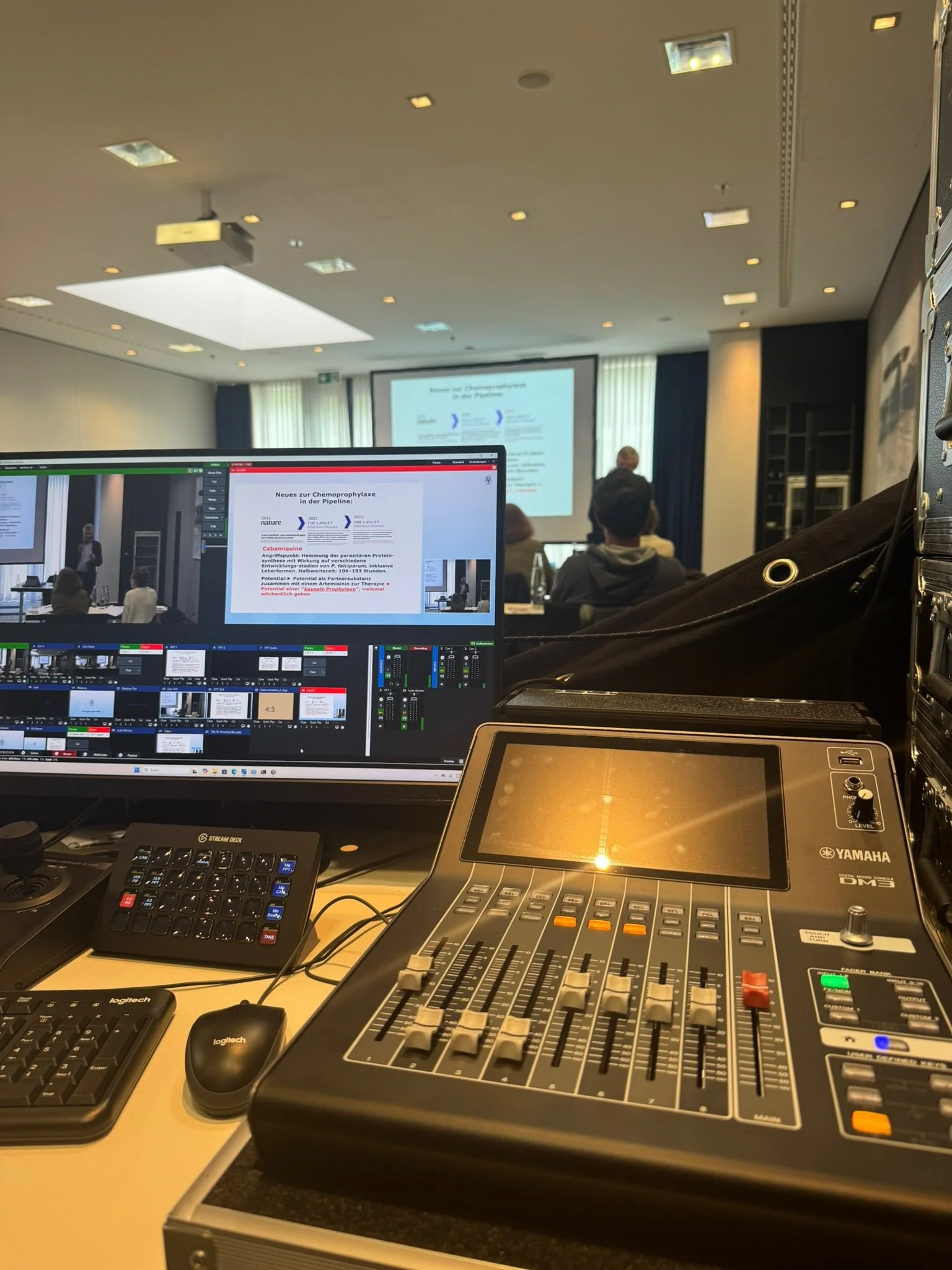 Aufnahme eines Konferenzraums mit mehreren Personen, einer Präsentation auf einer Leinwand und einer Fotoaufnahme von Technik, inklusive einem Laptop, einem Mischpult, einer Tastatur und einer Maus, für eine Präsentation oder einen Vortrag.