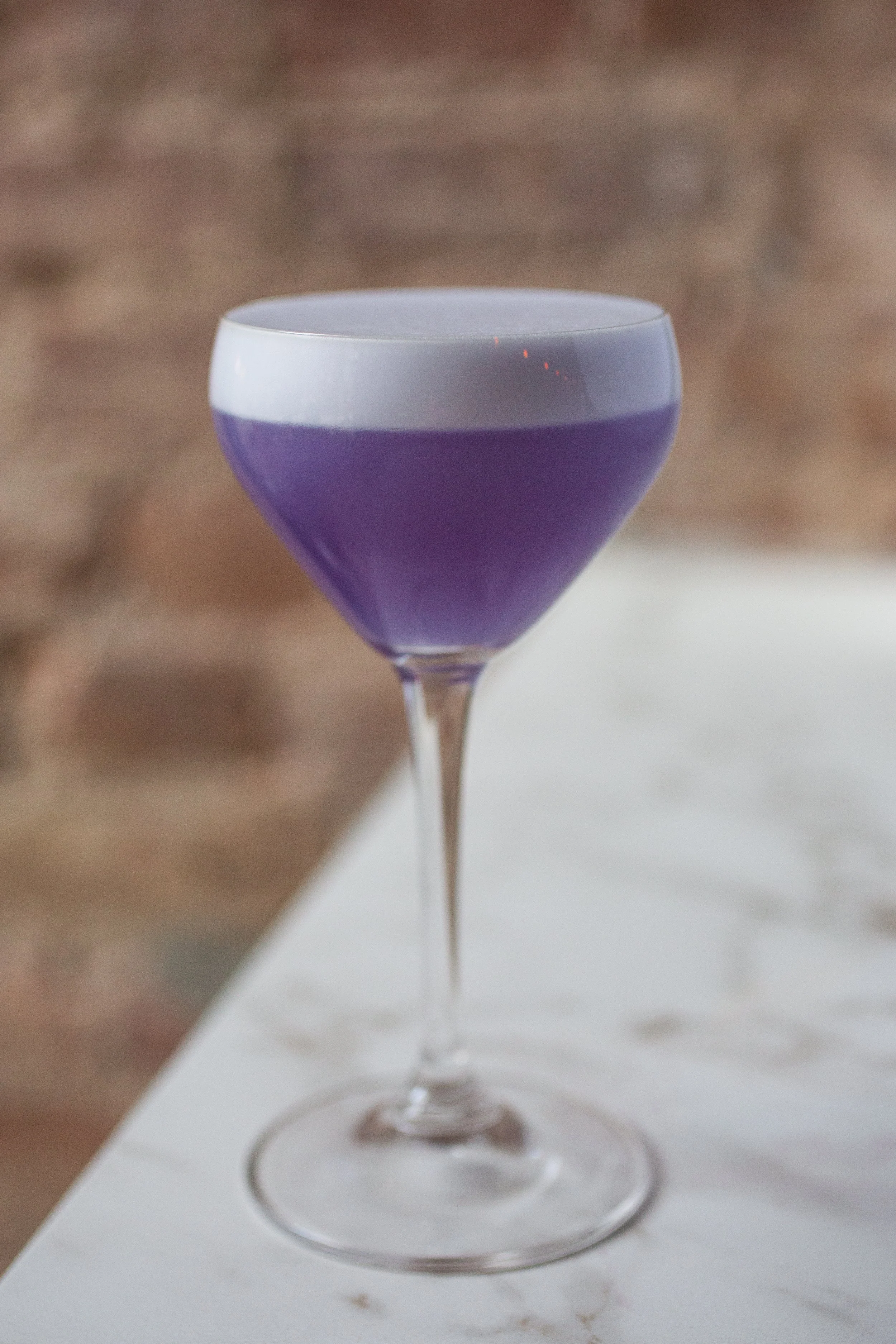 A cocktail glass with a purple drink topped with a gray foam layer, placed on a white surface with a blurred brick wall background.