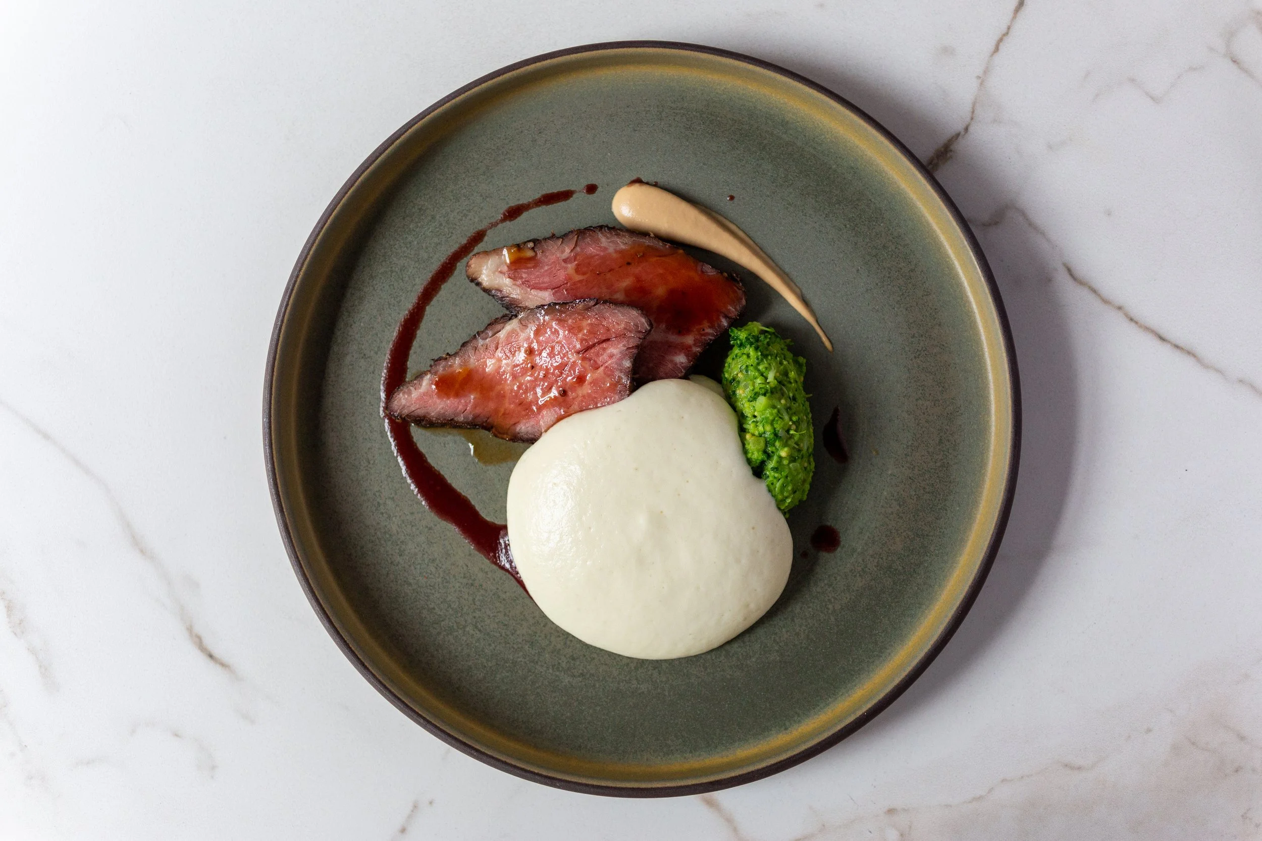 A dinner plate with slices of medium-rare beef, a dollop of white mashed potatoes, a piece of broccoli, a smear of beige sauce, and a drop of dark sauce on a green ceramic dish.