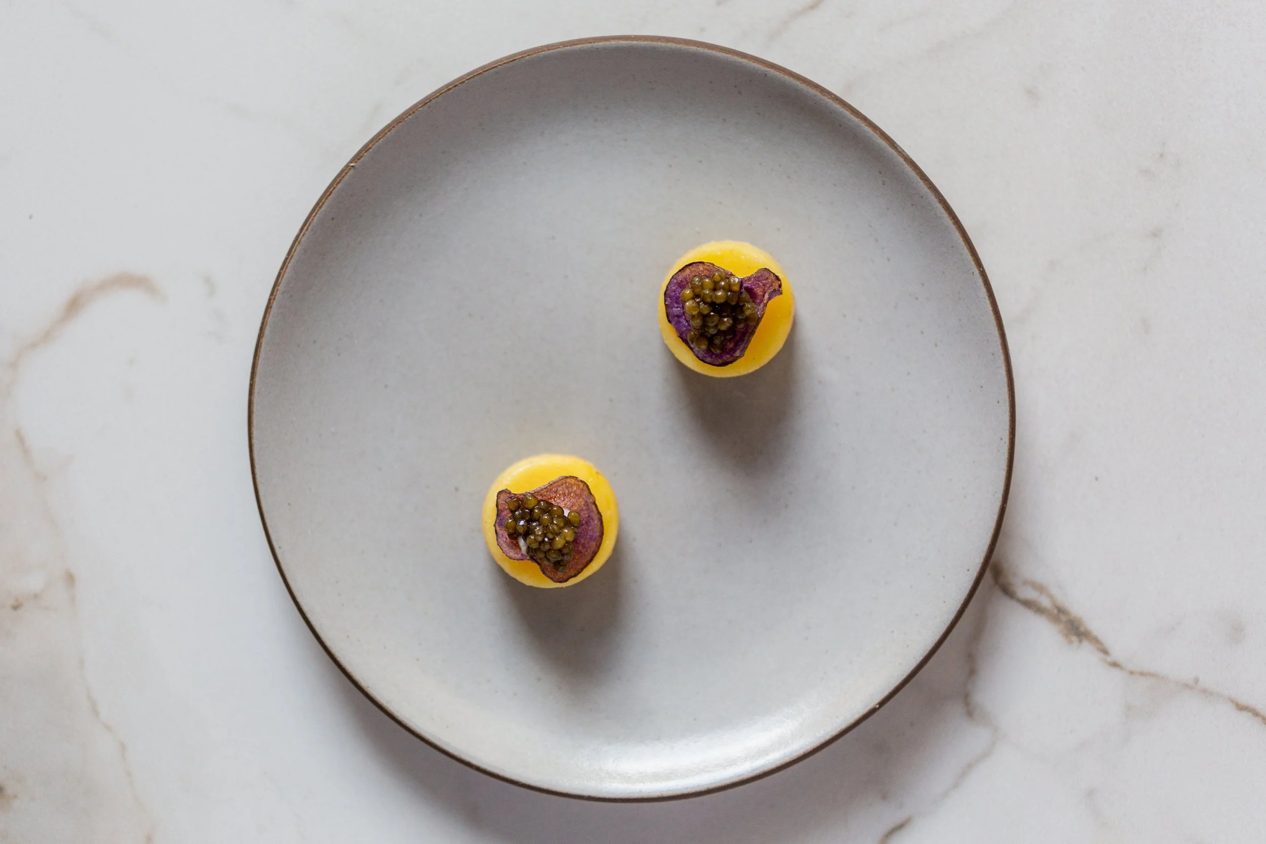 Two small, round yellow dishes topped with a purple garnish and black caviar, placed on a large gray plate on a white marble surface.