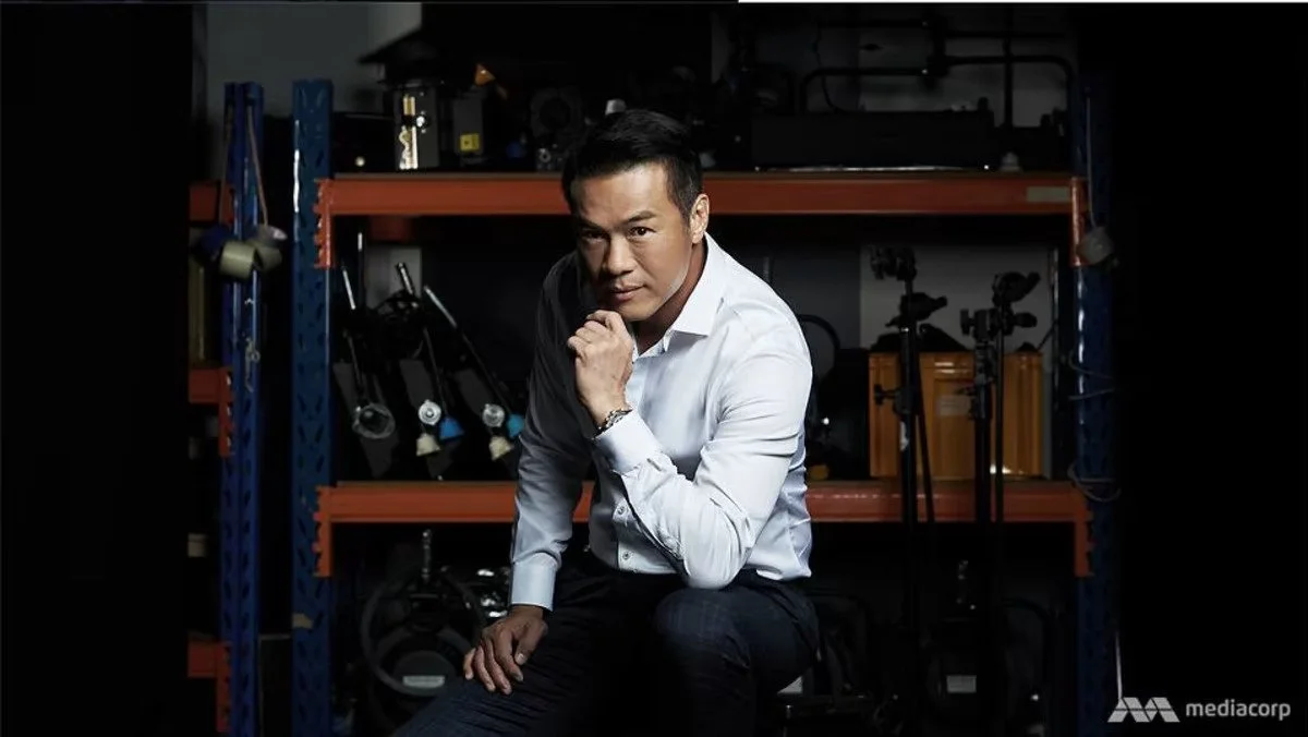 Man in white shirt sitting on a stool in a workshop, with camera equipment and tools on shelves behind him.