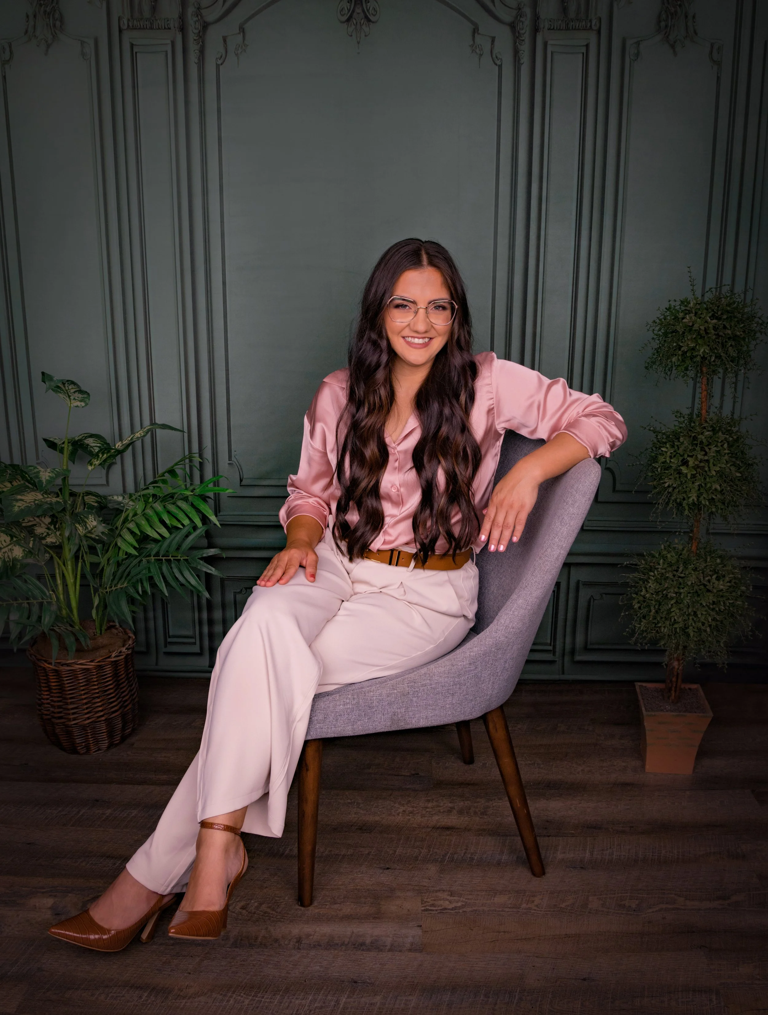 A woman with long dark wavy hair and glasses sitting on a grey chair, smiling, dressed in a pink silk blouse and beige pants, in a room with dark wood flooring, green plants, and ornate paneling on the wall.