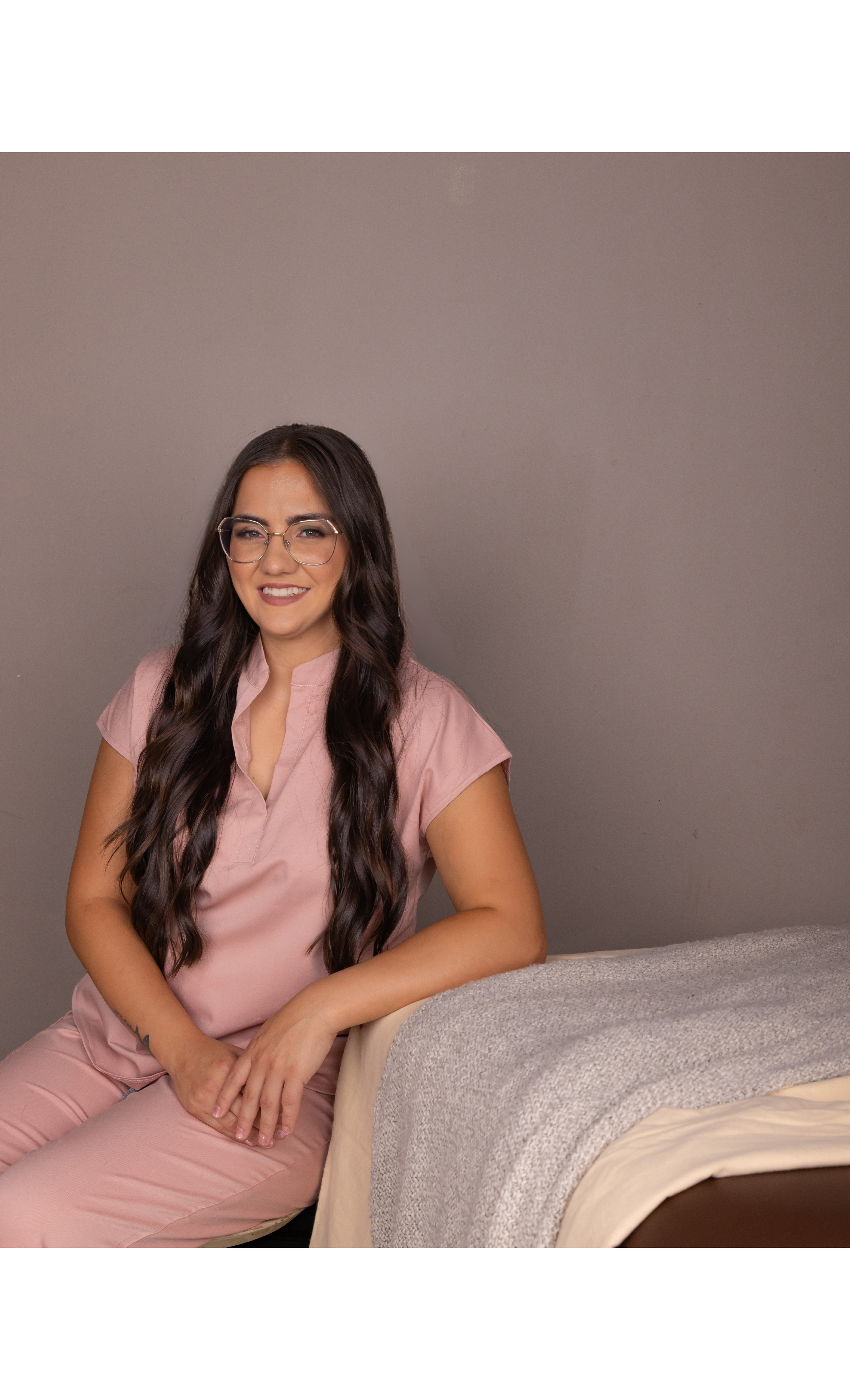 A woman with long dark wavy hair and glasses wearing a pink blouse sitting next to a bed with a gray blanket.