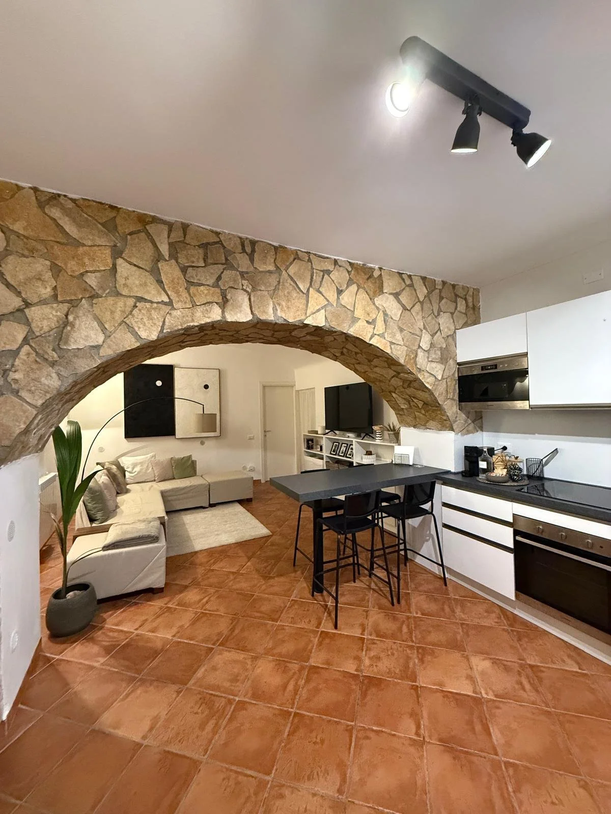 Interno di un soggiorno e cucina con archi in pietra, pavimenti in terracotta, soggiorno con divano, televisore, cucina con piano di lavoro nero e armadietti bianchi, illuminazione moderna