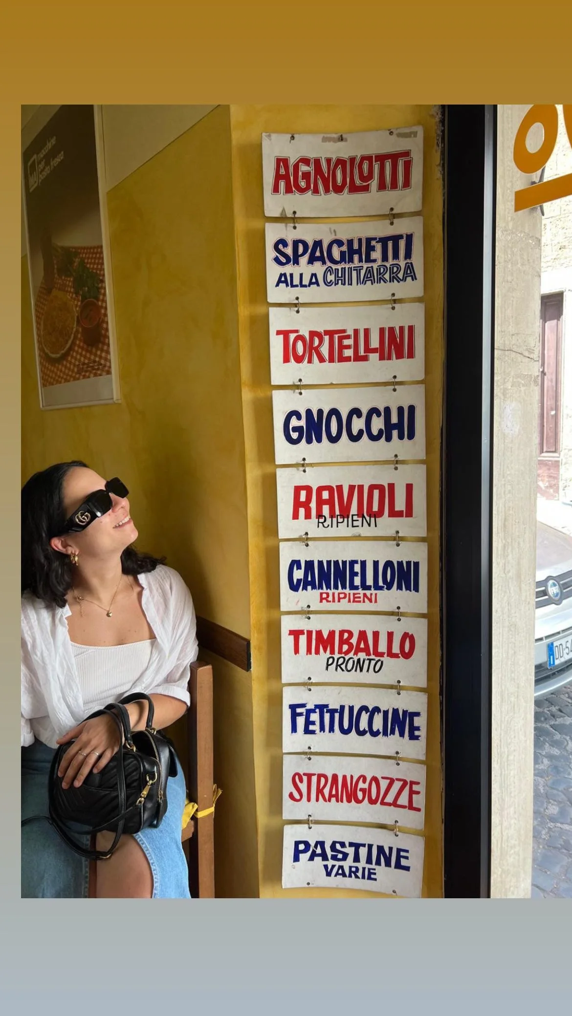 Un cartello di un ristorante con vari tipi di pasta e piatti italiani, tra cui spaghetti alla chitarra, tortellini, gnocchi, ravioli, cannelloni, timballo, fettuccine, strangoze e paste varie. Una donna seduta a fianco guarda sorridendo verso il pannello.