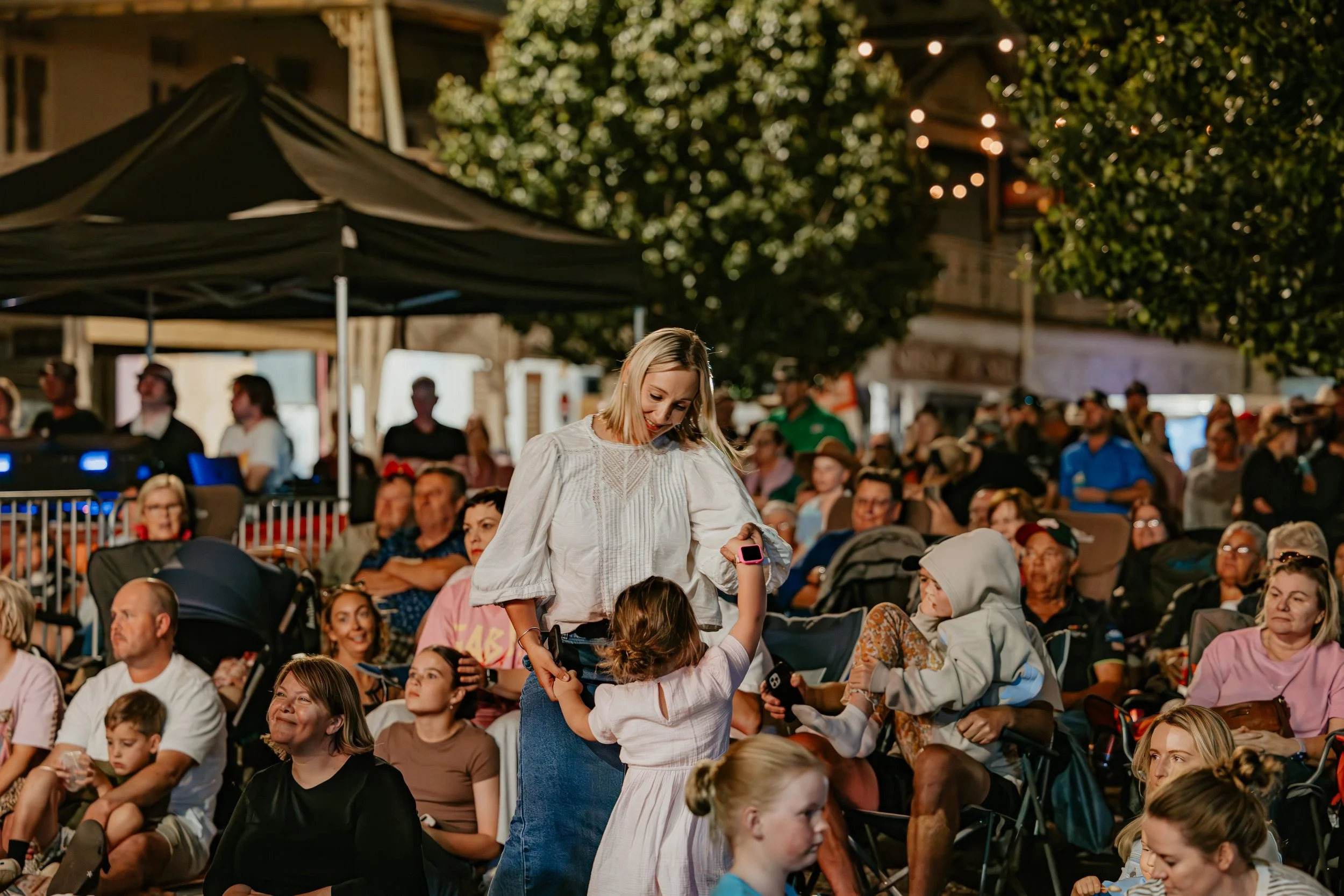 A woman is dancing and smiling with a young girl at an outdoor event during evening, surrounded by a seated crowd under trees and string lights.
