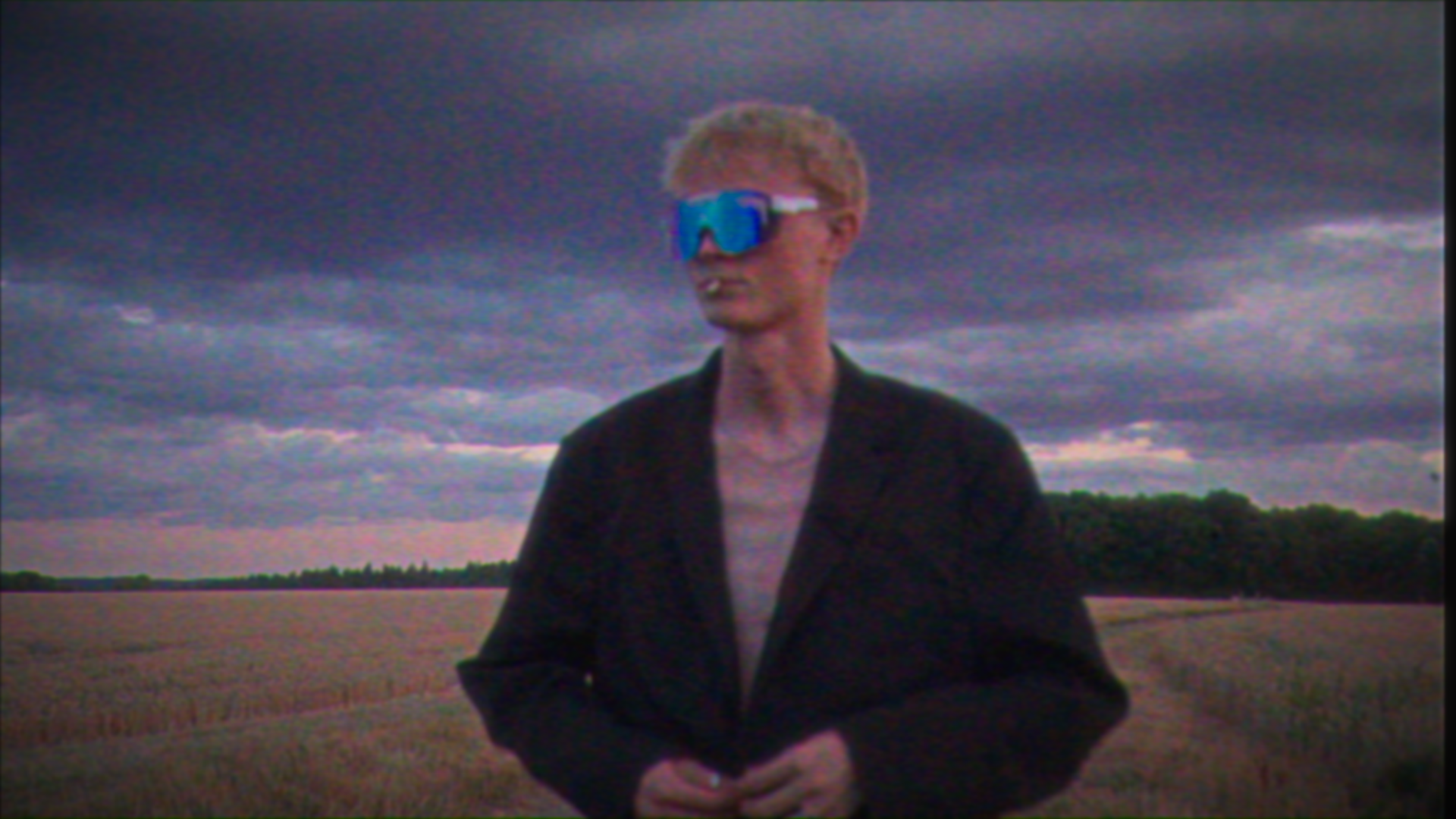 Person with blond hair wearing reflective sunglasses and a black jacket standing outdoors in a field under a cloudy sky.