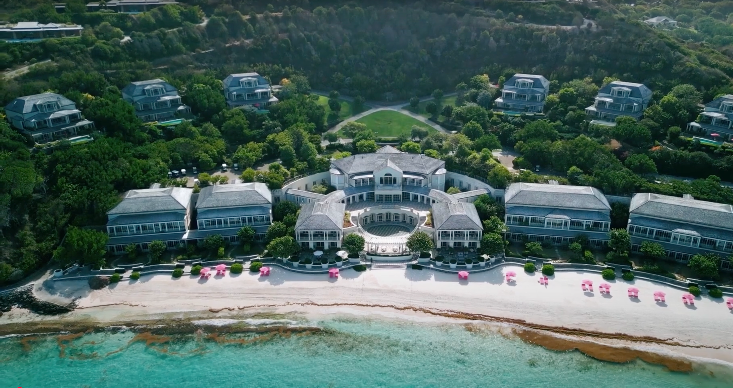 Aerial view of a beachfront resort with multiple large buildings, a curved central structure, and pink umbrellas on a sandy beach, overlooking turquoise water.