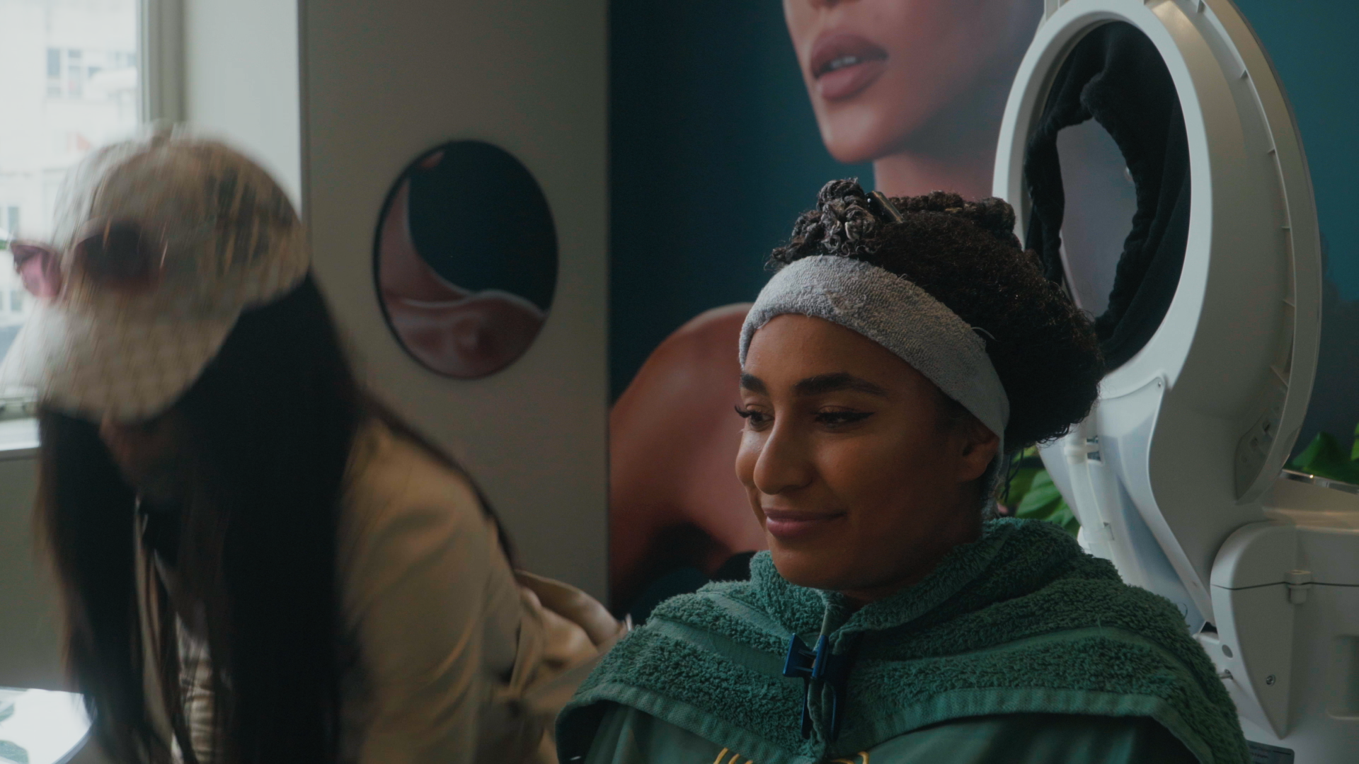A woman undergoing a medical imaging or dental procedure, with a healthcare professional beside her. The woman is sitting calmly, wearing a towel around her shoulders and a headband, with an MRI or similar machine in the background and a large advertisement of a woman's face on the wall.
