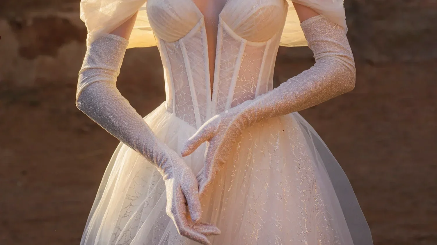 Close-up of a woman in a white wedding dress with long gloves, standing outdoors during sunset.