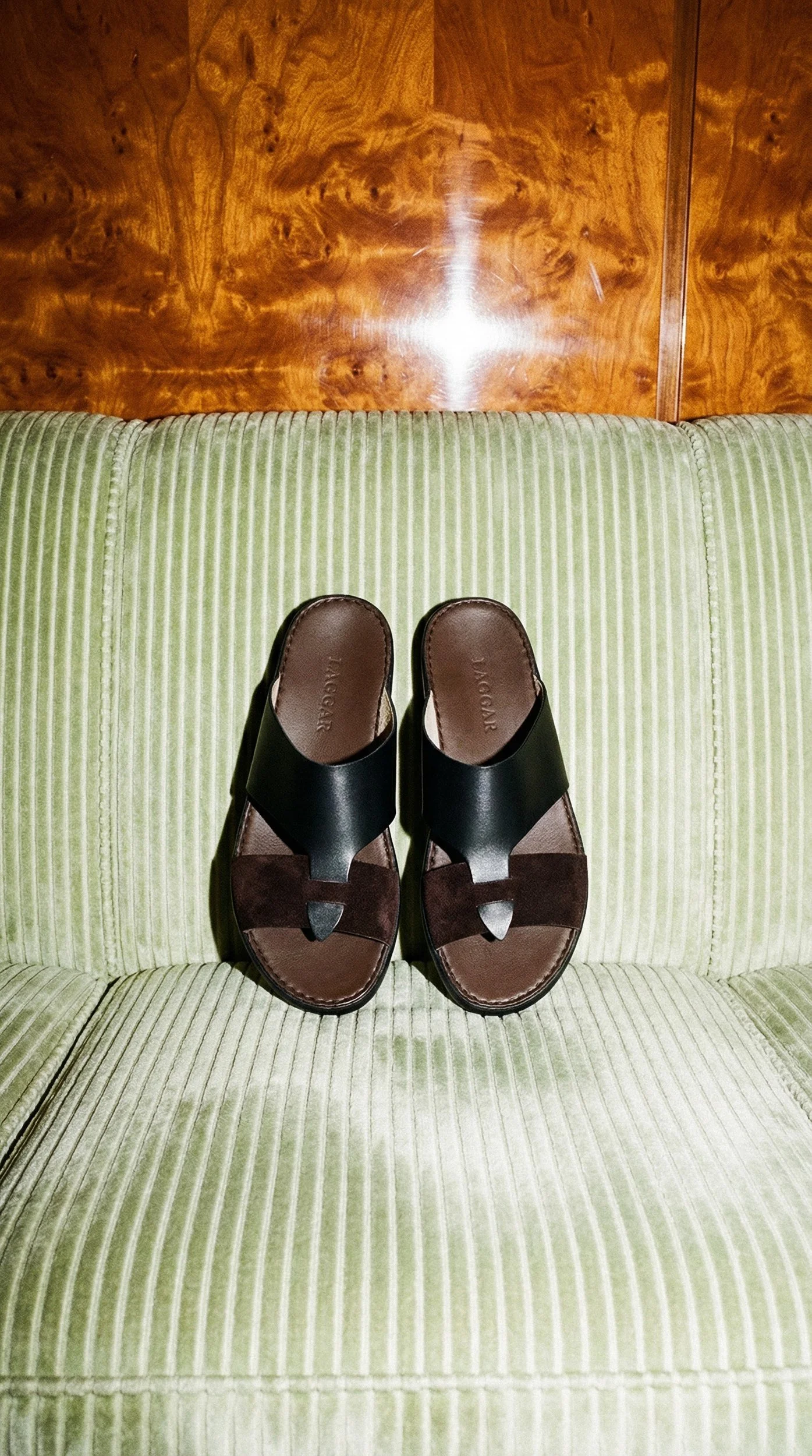 Pair of brown and black sandals placed on a light green striped couch with a wooden wall background.