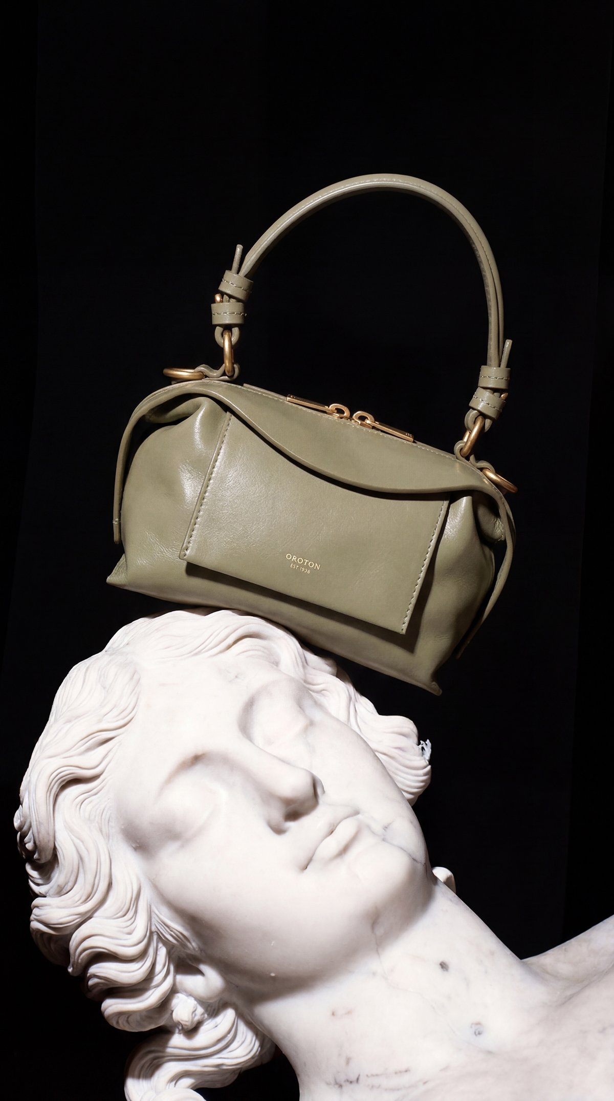 A white marble sculpture of a woman's head with closed eyes, relaxed expression, and flowing hair, positioned against a black background. Resting on the sculpture's head is a stylish green handbag with gold hardware and a front pocket.