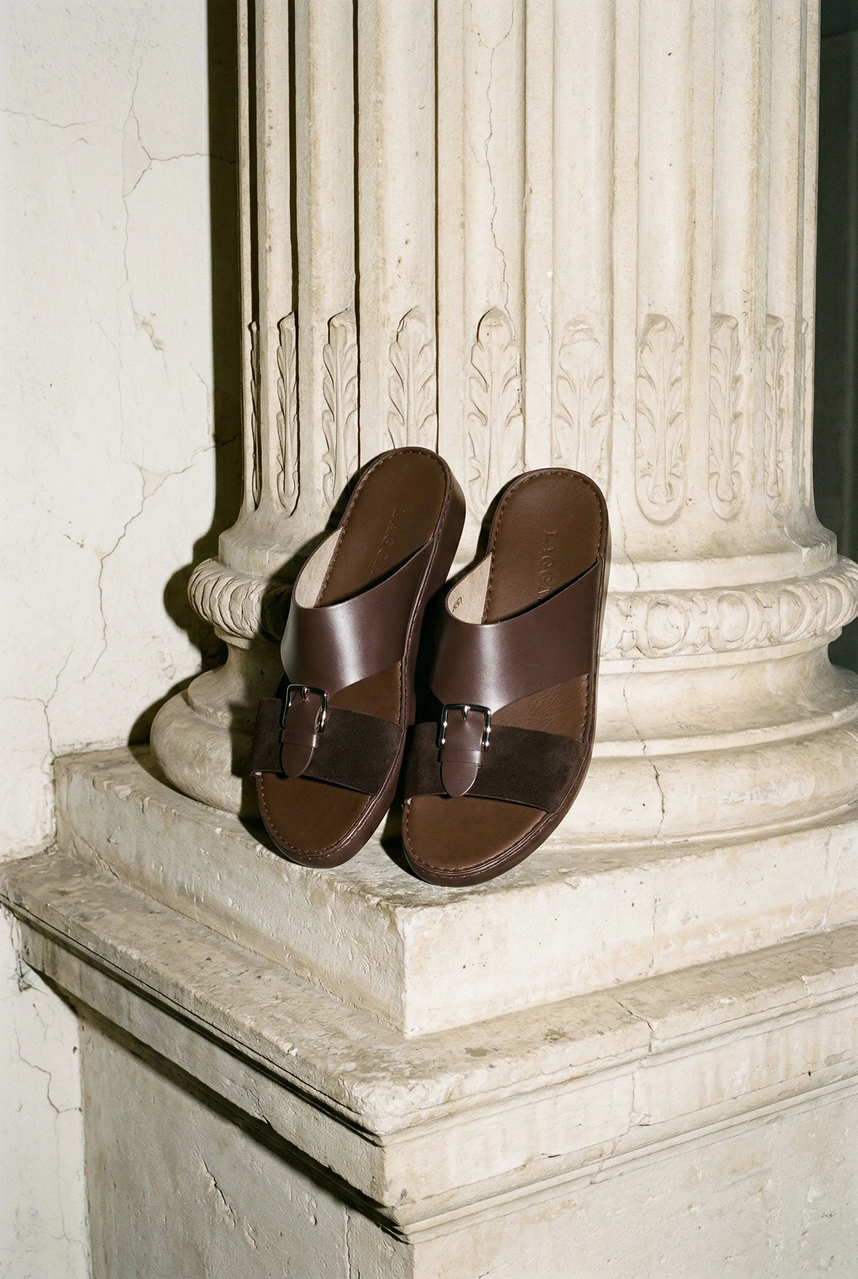Brown slide sandals placed on a stone pedestal in front of a decorative carved column.