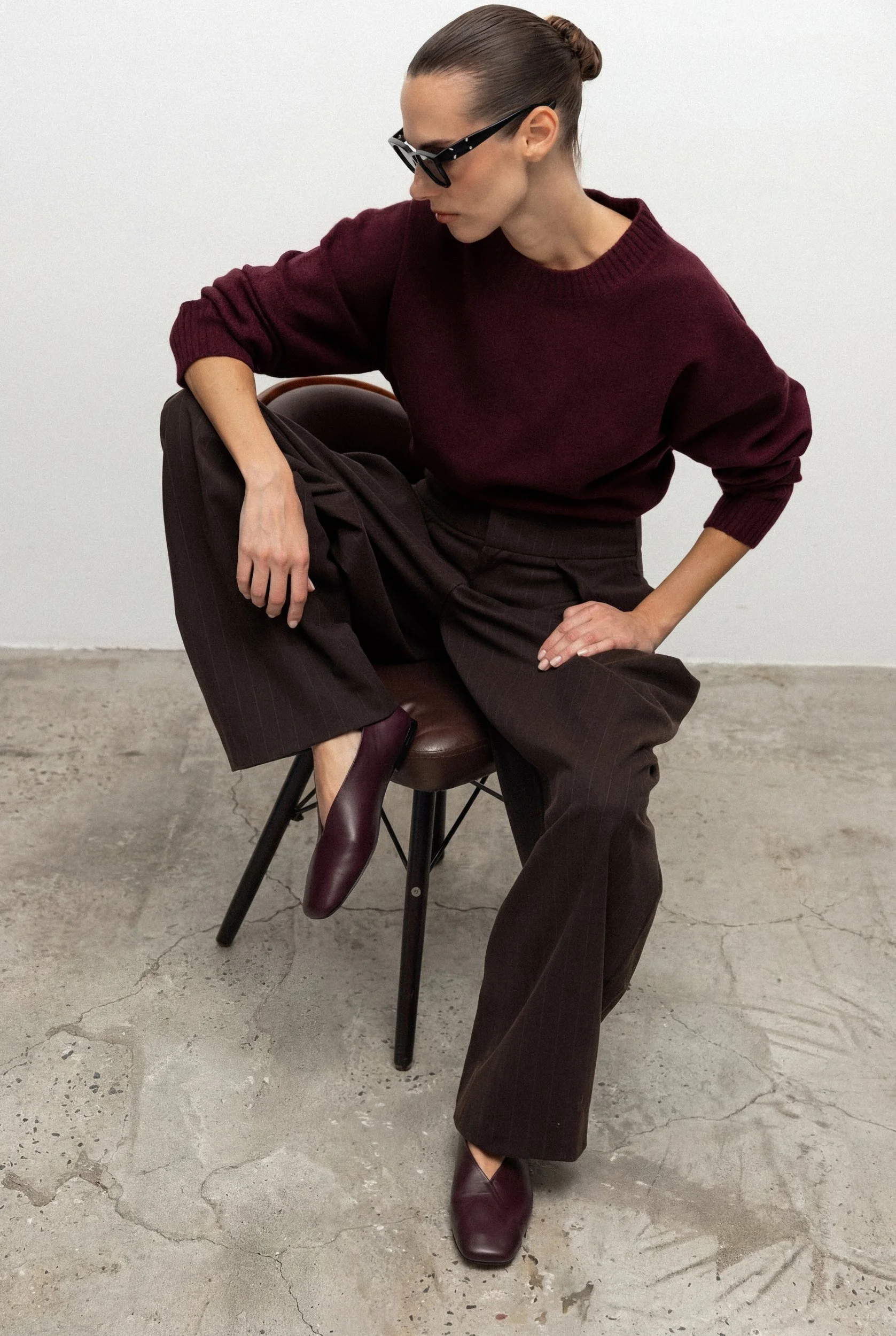 A woman wearing a burgundy sweater, dark brown wide-leg pants, maroon ankle boots, and black sunglasses is sitting on a chair in a minimal environment with a plain light background and a cracked concrete floor.