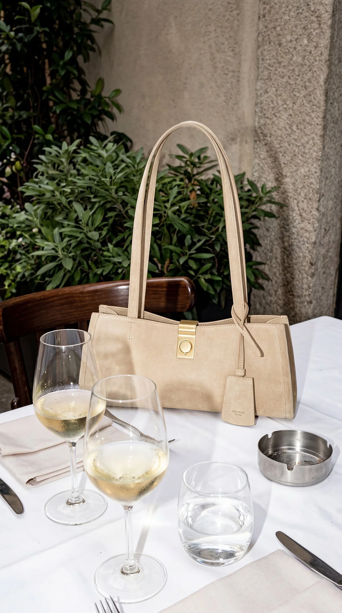 A beige handbag with gold hardware placed on a white tablecloth at an outdoor dining setting, with wine glasses, water glasses, a napkin, and utensils, and green plants and stone wall in the background.